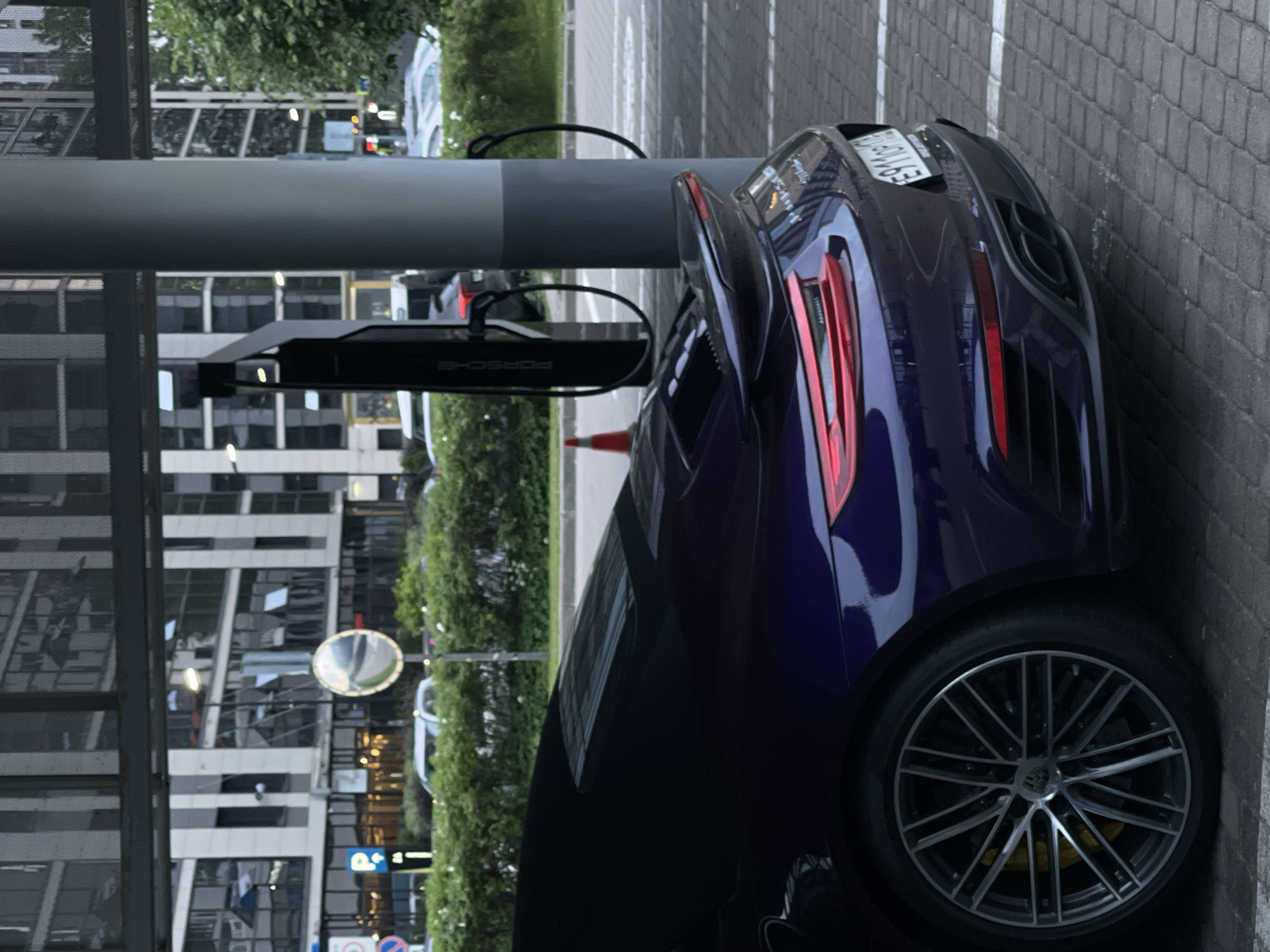 A dark purple Porsche 911 is parked at a charging station. The car is viewed from the rear passenger side. The vehicle has a large rear spoiler and prominent exhaust pipes. A charging cable is visible attached to a charging station with the Porsche logo. In the background, modern buildings and lush green bushes are seen. The ground is paved with interlocking bricks. The scene appears to be in an urban setting, possibly a parking lot or a dedicated EV charging area.