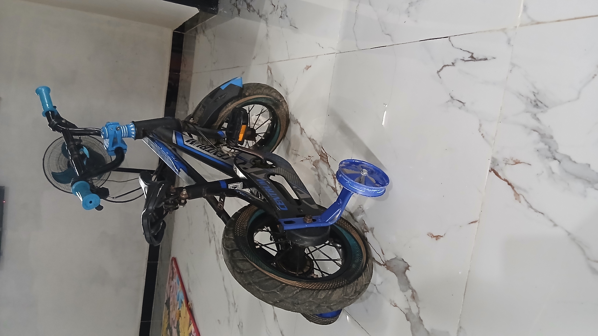 A children's bicycle, predominantly black and blue, with training wheels is parked on a tiled floor. The bicycle features a black frame with blue accents and graphics. It has blue handlebar grips and blue seat. The training wheels are also blue. The floor is tiled with a white and brown marble pattern. In the background, a black oscillating fan is visible against a light-colored wall. A colorful children's play mat is partially visible on the left side of the image. The scene appears to be indoors, likely in a residential setting. There are no people present in the image.