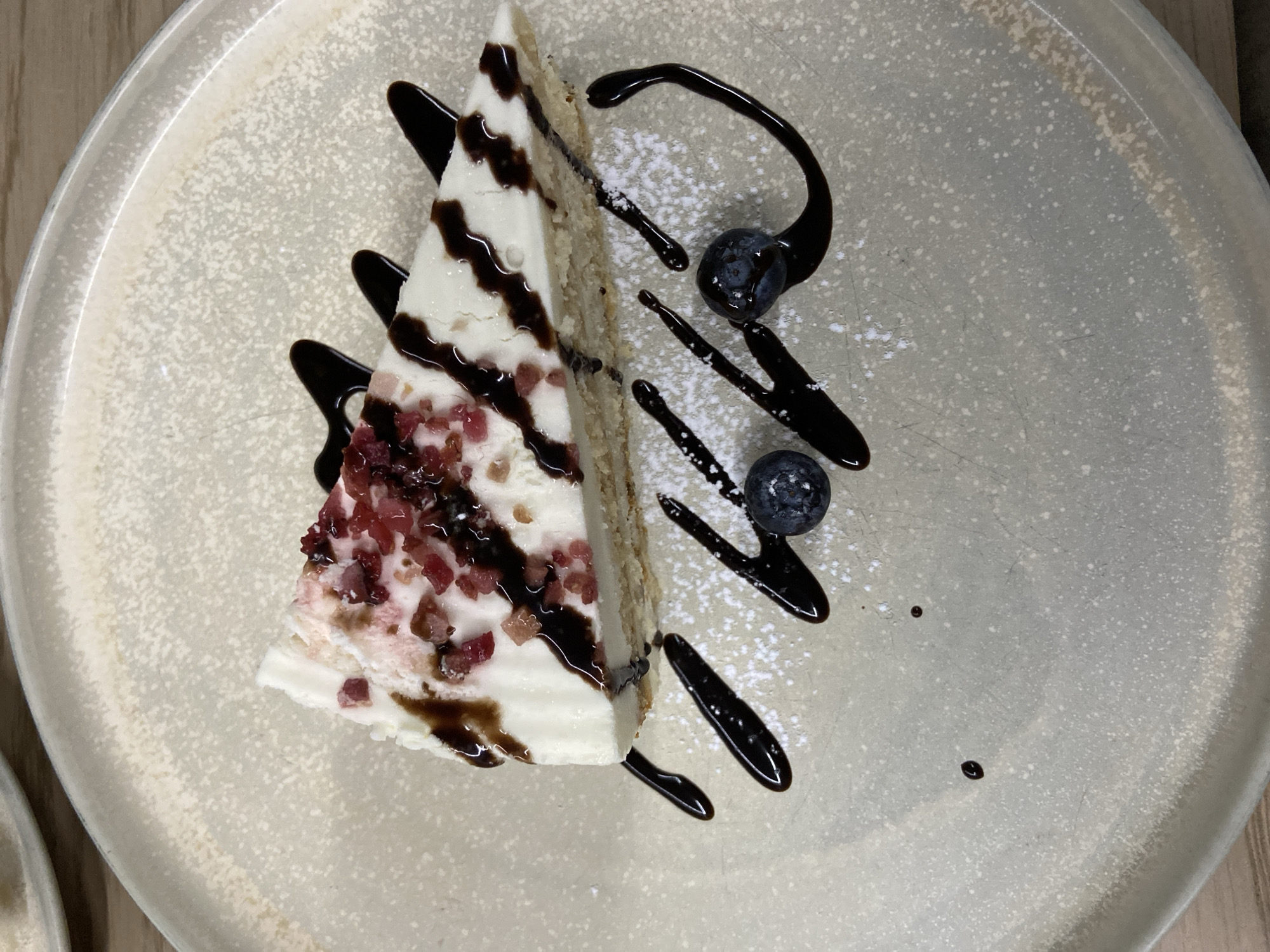 A slice of cake with white frosting and crumbled pink topping sits on a speckled gray plate, adorned with a drizzle of dark chocolate sauce. Two blueberries and a dusting of powdered sugar are also present on the plate. The background is slightly out of focus, showing a hint of a wooden surface. This image was taken in Tuymazy, Russia.