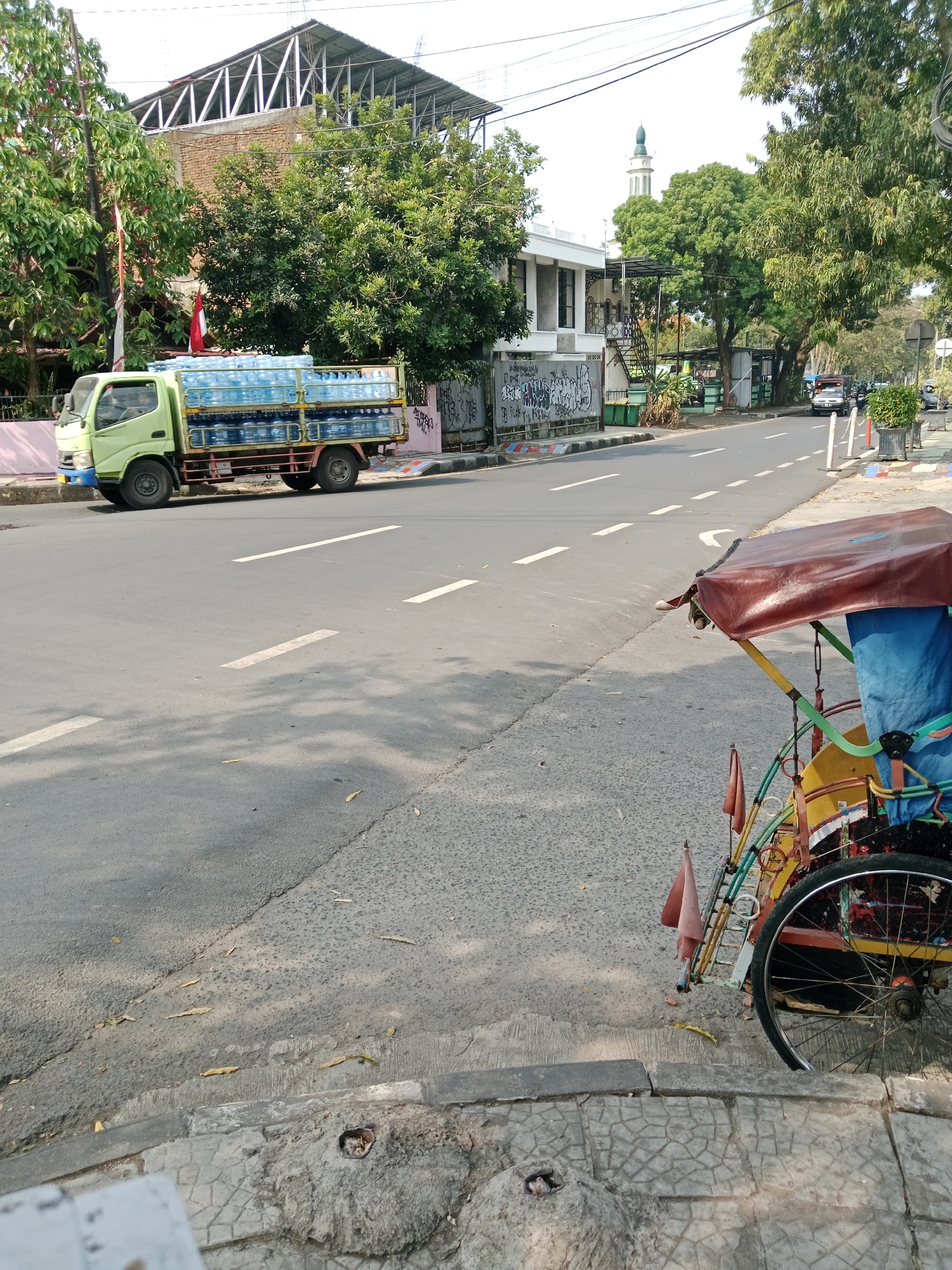 The image captures a street scene in Ciamis, Indonesia. In the foreground, a colorful, three-wheeled vehicle, likely a pedicab or becak, is parked on a concrete sidewalk. The vehicle has a maroon canopy and a blue fabric covering. A light green truck is parked on the right side of the road, laden with multiple stacks of large, clear plastic bottles filled with water. The truck is positioned near a building with a modern facade and a red and white Indonesian flag attached to a pole. Across the street, a mosque with a prominent minaret is visible among lush green trees. The road is paved and marked with white lane lines. The environment appears to be a sunny day, with shadows cast by trees on the road. The street is bordered by sidewalks and vegetation.