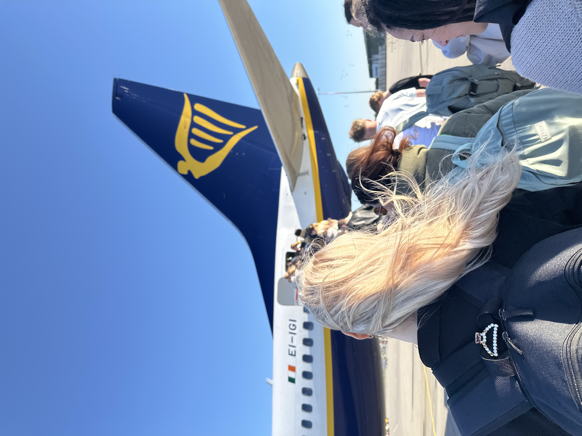 This image captures an outdoor scene at an airport, likely on the tarmac, bathed in bright daylight under a clear blue sky. A group of people are viewed from behind, appearing to queue or walk towards the rear entrance of a large passenger airplane, indicating a boarding process is underway.
The dominant object is a Ryanair aircraft, identifiable by its distinct dark blue tail fin featuring the airline's yellow harp logo. The plane's fuselage is white with a yellow stripe and blue livery. Clearly visible on the side of the fuselage is the aircraft registration "EI-IGI" positioned next to a small Irish flag, confirming its Irish registration. The people are moving towards an open door at the back of the aircraft.
In the foreground, closest to the camera, is the back of a person with long, blonde or light brown hair, wearing a dark top and a black backpack. A decorative hair clip, possibly made of white beads or pearls, is attached to a strap of their backpack. Further into the frame, other individuals with varied hair colors (including dark, brown, and red-brown) are seen, also from behind. They are dressed in casual clothing, such as sweatshirts and jackets, and some are carrying backpacks, including a prominent light green/teal one.
The bright sunlight suggests it is daytime, with clear weather conditions. The scene depicts passengers boarding a flight, likely a low-cost carrier, at an airport. Given the provided context, the event is taking place in Weeze, Germany.