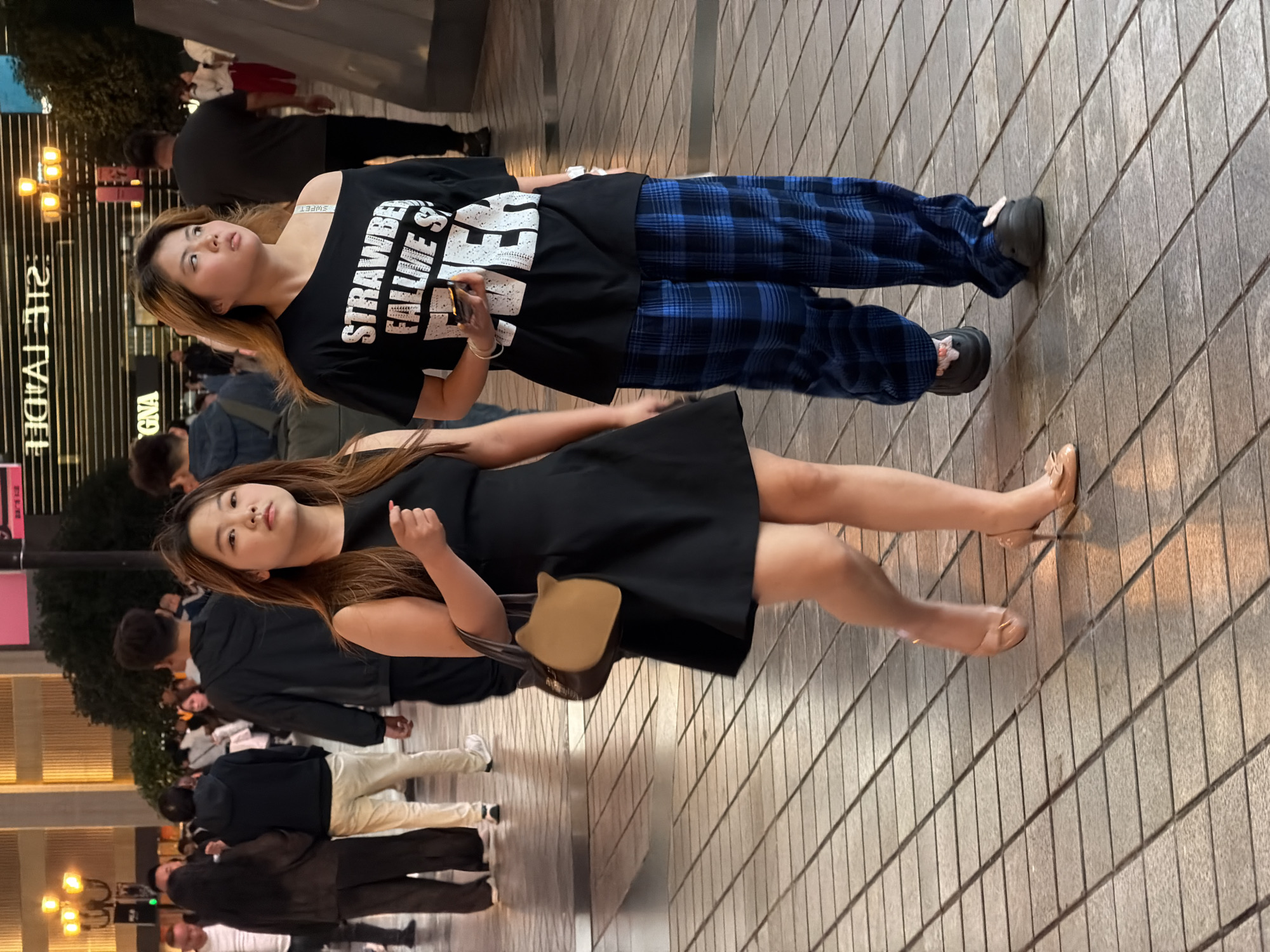Two young women are walking on a paved sidewalk in Yuzhong District, China. The woman on the left is wearing a black sleeveless dress and nude high heels. She is carrying a tan handbag with a cat-ear design. The woman on the right is wearing a black off-the-shoulder t-shirt with white lettering that reads "STRAWBERRY FALL ME FOREVER" and blue plaid pants. She is wearing black chunky shoes. In the background, other pedestrians can be seen, as well as illuminated buildings and streetlights, suggesting it is evening or night. The pavement is made of square tiles.