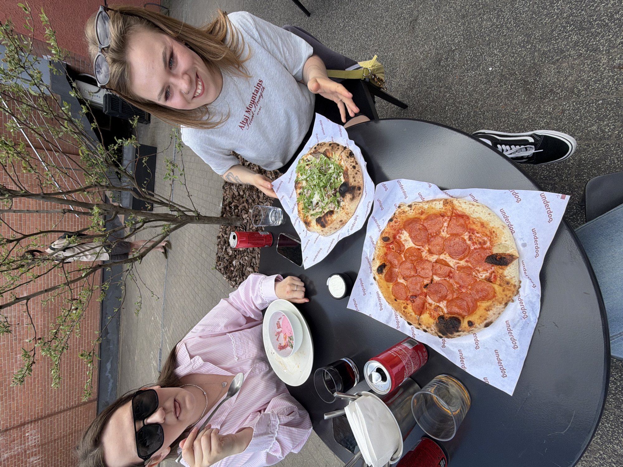 Here is a forensic description of the image.
The image shows an outdoor scene in what appears to be a casual dining setting. There are two young women at a black round table, apparently enjoying a meal. One woman, positioned in the upper part of the frame, is smiling and gesturing towards a pizza in front of her. She is wearing a gray t-shirt with the words "Altai Mountains" and has sunglasses perched on her head. The other woman is in the lower part of the frame, wearing sunglasses and a pink striped shirt. She is holding a spoon and has a plate of what looks like dessert in front of her.
On the table, there are two pizzas on paper printed with the word "underdog," indicating the restaurant. One is a pepperoni pizza, and the other has arugula and other toppings. There are also cans of soda, glasses, a small container, and a white purse or wallet on the table. The table sits on a paved surface. A small tree is positioned to the side. A person's shoe is partially visible under the table.
The image doesn't provide specific clues to determine the location of the scene within Minsk, Belarus.