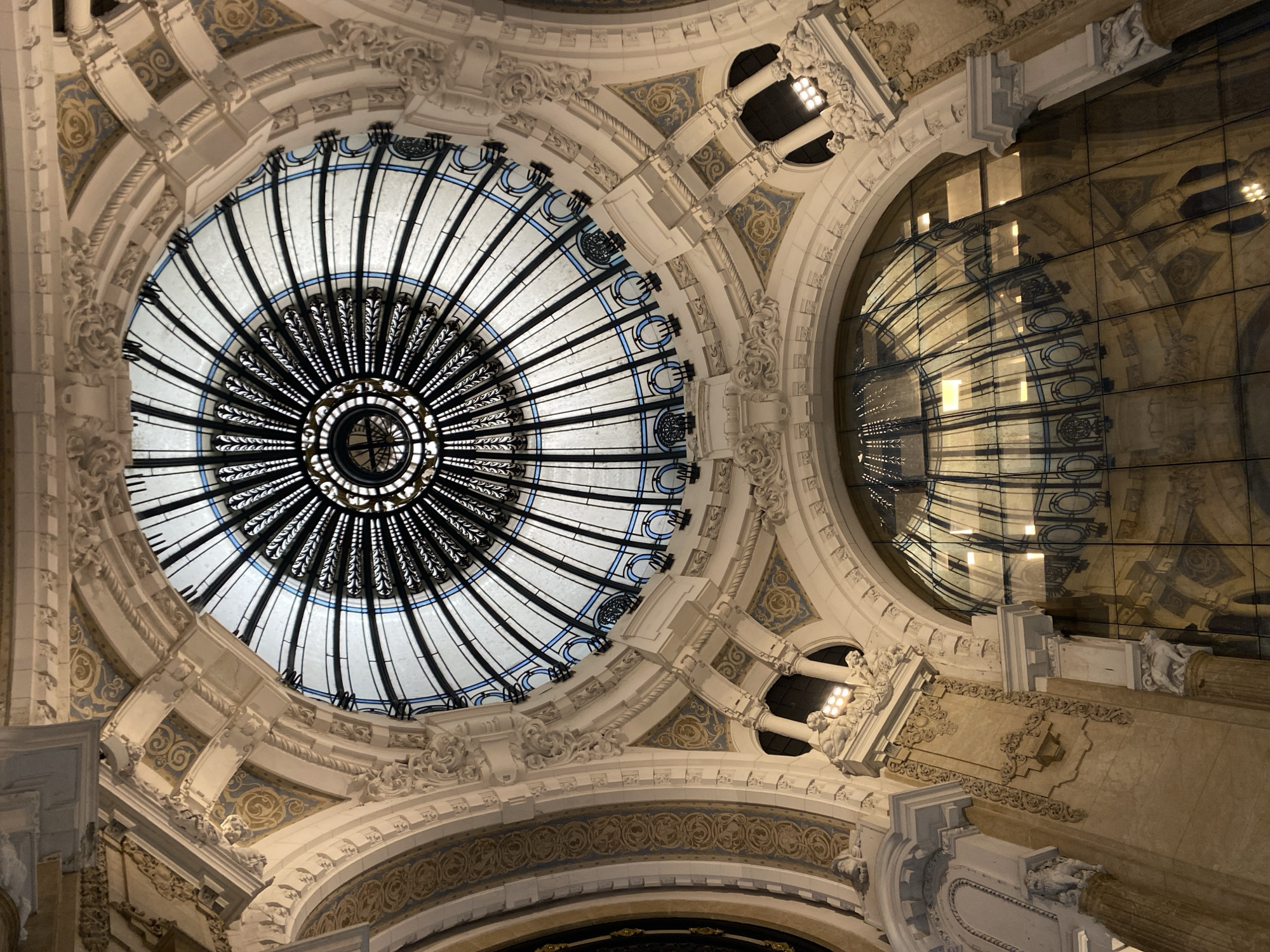 The image shows the interior of a building, specifically the dome or ceiling area. The style appears to be ornate, with intricate architectural details and decorative elements. A central circular stained-glass skylight dominates the composition. The skylight has a complex pattern with radiating lines and a central circular motif. The surrounding architecture features classical-style ornamentation, including moldings, carvings, and possibly statues. To the right of the skylight is a curved glass or reflective surface, likely part of the building's structure or design. The image is taken from a low angle, looking upwards toward the ceiling.
This is a view from inside Galerías Pacífico, a shopping mall located in the city of Buenos Aires, Argentina. The architecture is a prime example of Beaux-Arts style.