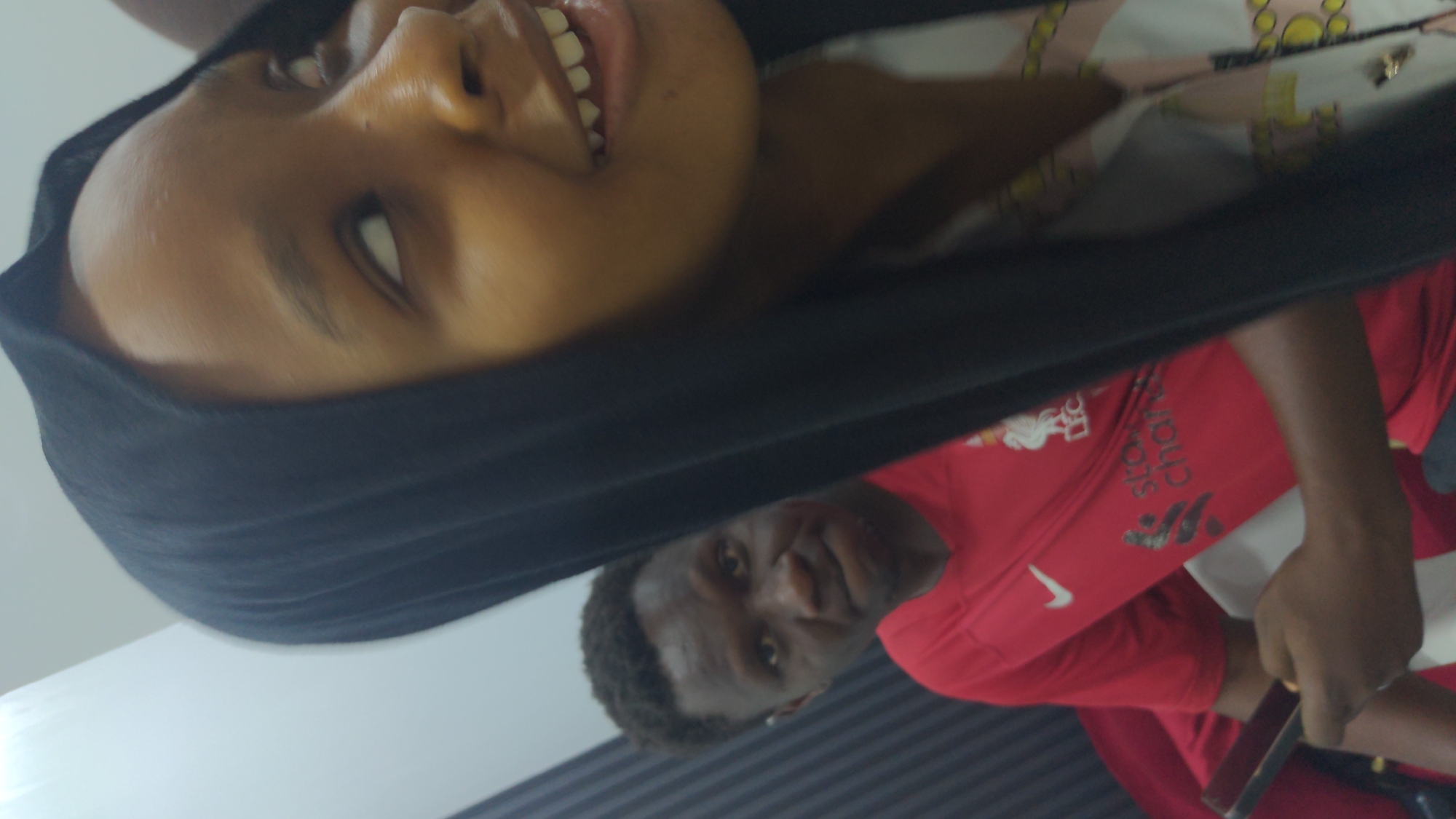The image, taken in Abuja, Nigeria, features two individuals indoors. In the foreground, a woman with dark skin is shown from the head to the upper torso, wearing a dark head covering and a light-colored, patterned top. She is smiling broadly, looking upwards and to her left. Behind her and to the right, partially obscured, is a man with dark skin, visible from the head to the upper torso. He is wearing a red t-shirt and looks towards the viewer with a neutral to slightly smiling expression. The red t-shirt displays a white Nike swoosh, the Liverpool Football Club (LFC) crest, and the text "Standard Chartered." The man appears to be holding a small device or phone in his right hand. The background includes a plain light-colored surface and a wall with horizontal dark and light stripes. The lighting suggests an indoor setting during the daytime.