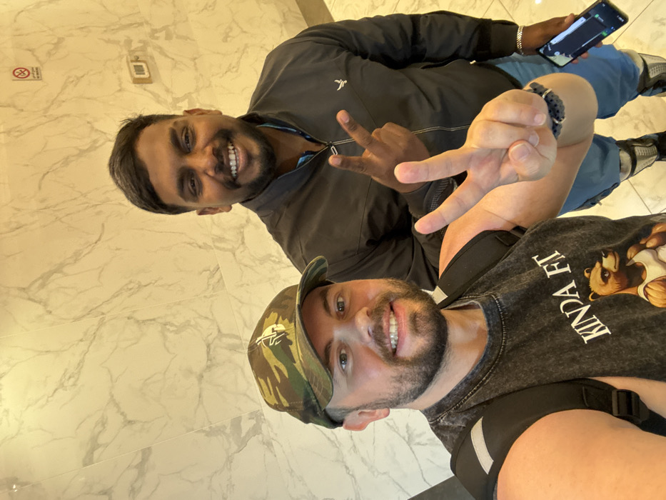 Two men are posing for a selfie. One man is wearing a black jacket, blue jeans, and white sneakers. He's holding a phone and making a hand gesture. The other man is wearing a camouflage hat and a black t-shirt. Both men are smiling. The background is a marble wall with a "no smoking" sign.