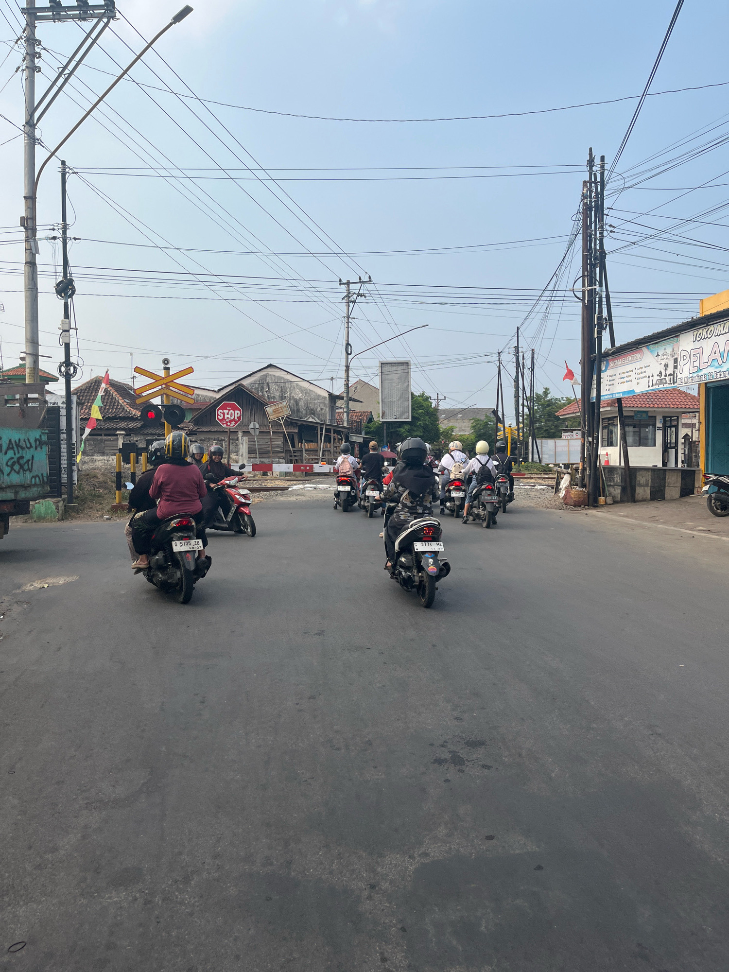The image captures a street scene in Karangasem Selatan, Indonesia, featuring a procession of motorbikes navigating an asphalt road under a clear sky. The road is lined with various structures, including utility poles with a complex network of wires, and buildings. On the right side of the image, a storefront with a yellow awning and signage that reads "TOKO" and "PELANG" is visible, along with a white building featuring a red roof and windows. A red and white Indonesian flag flies prominently on a pole near the storefront. To the left, a green truck with graffiti on its side is parked. A key element in the scene is a railway crossing with a yellow crossing signal and a red "STOP" sign. Several individuals are riding on the motorbikes, most wearing helmets for safety. The overall atmosphere suggests a typical day in a bustling Indonesian town, with traffic and everyday life unfolding on the street.