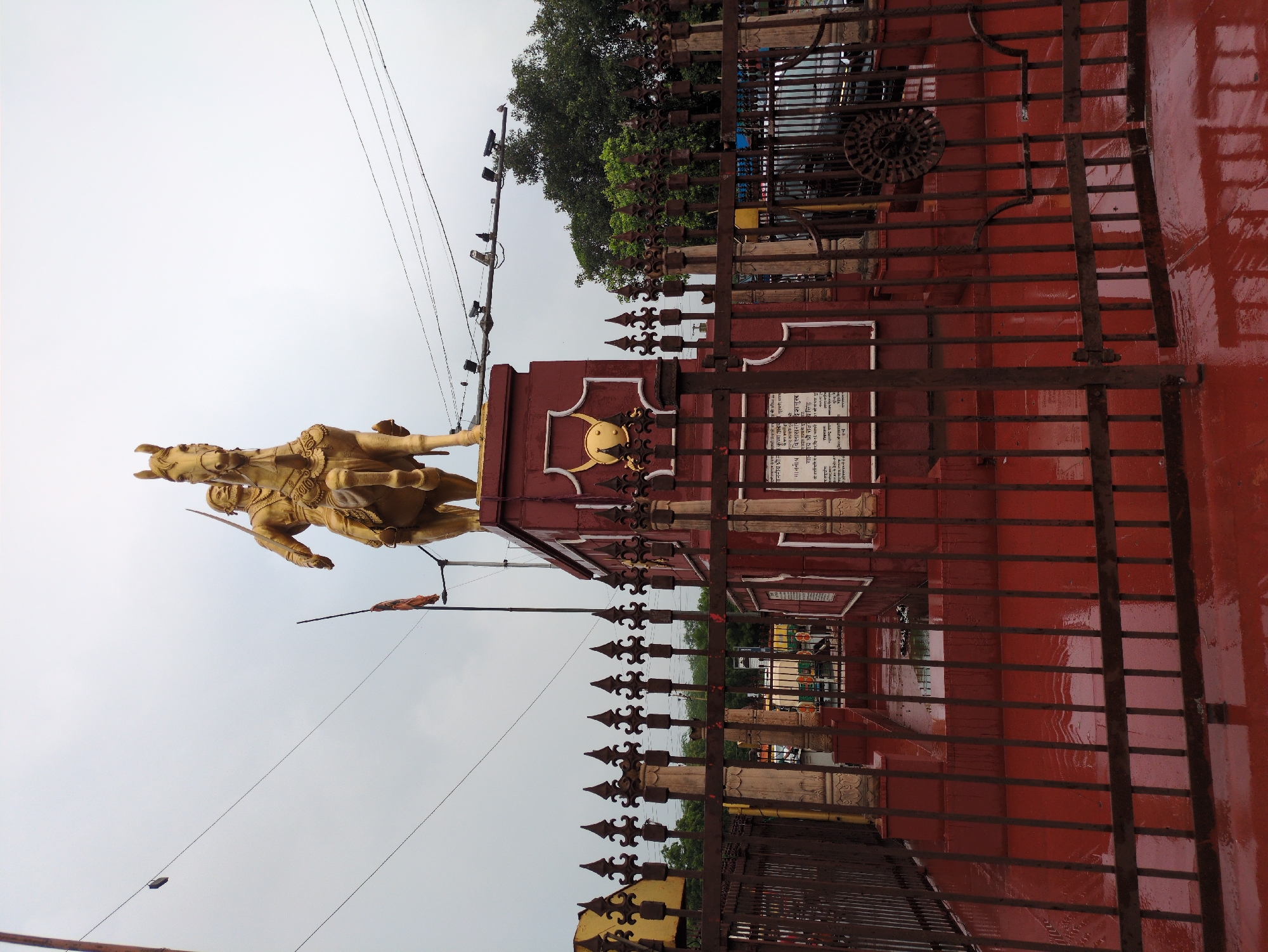 A golden statue of a man on horseback, holding a sword, stands atop a red, ornate pedestal. The statue is located in Agra, India, and is surrounded by a decorative metal fence. The ground in front of the fence is wet and reflects the sky, suggesting it has recently rained. The sky is overcast, indicating a cloudy day. Behind the statue and fence, trees and a building are visible, as well as a few vehicles. A flag is planted next to the statue. The scene appears to be a public memorial or landmark.