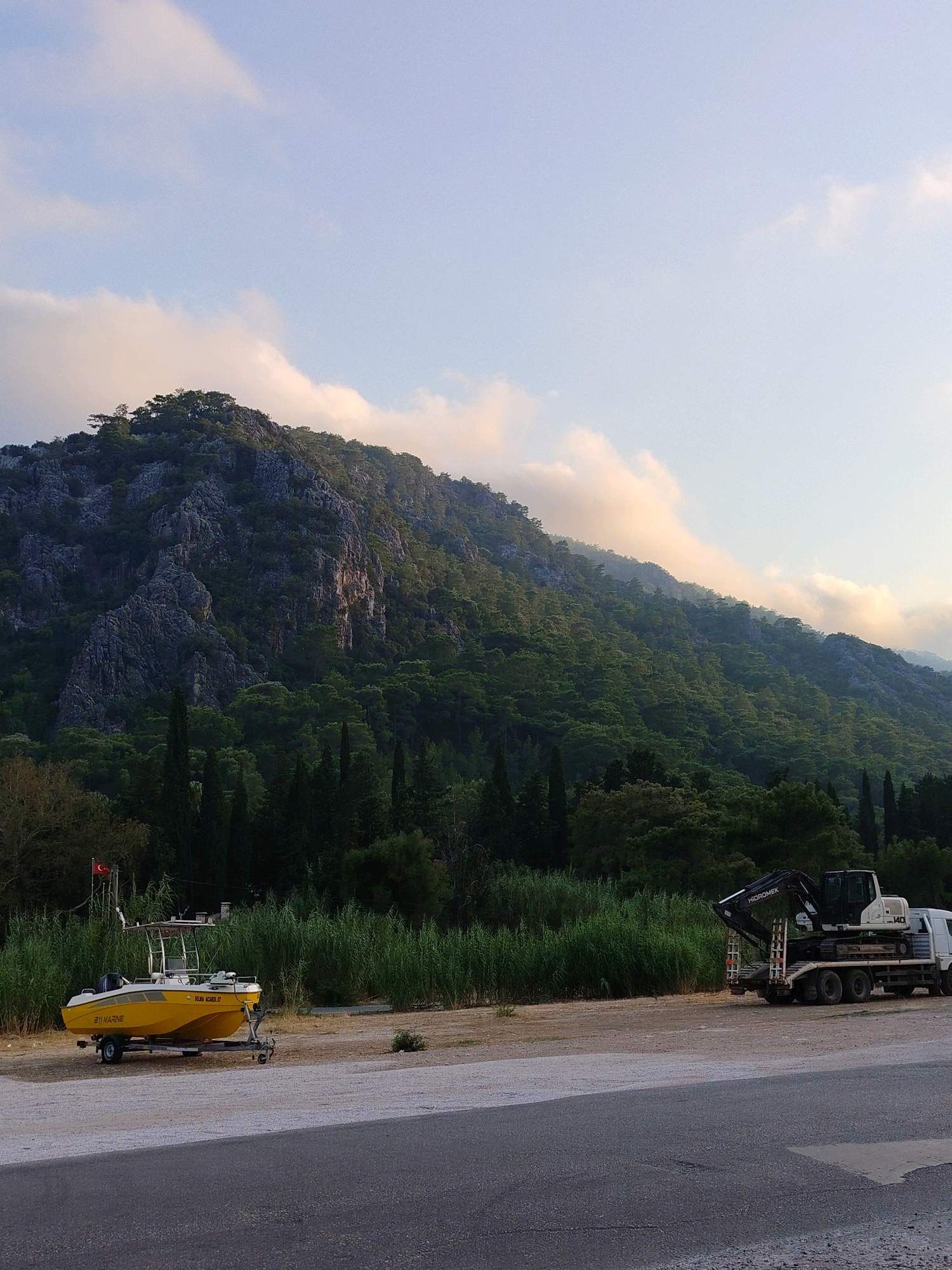 The image depicts a scenic view of a mountainous landscape with lush green trees and foliage in the foreground. A bright yellow boat on a trailer is parked on a dirt and gravel area, accompanied by a large white truck carrying a dark excavator. A Turkish flag is visible on a pole near the boat. The sky is a pale blue with scattered clouds, some illuminated by sunlight. The scene appears to be a roadside or parking area with a natural backdrop. The location is identified as Konyaaltı, Turkey, a district known for its coastline and mountainous terrain.