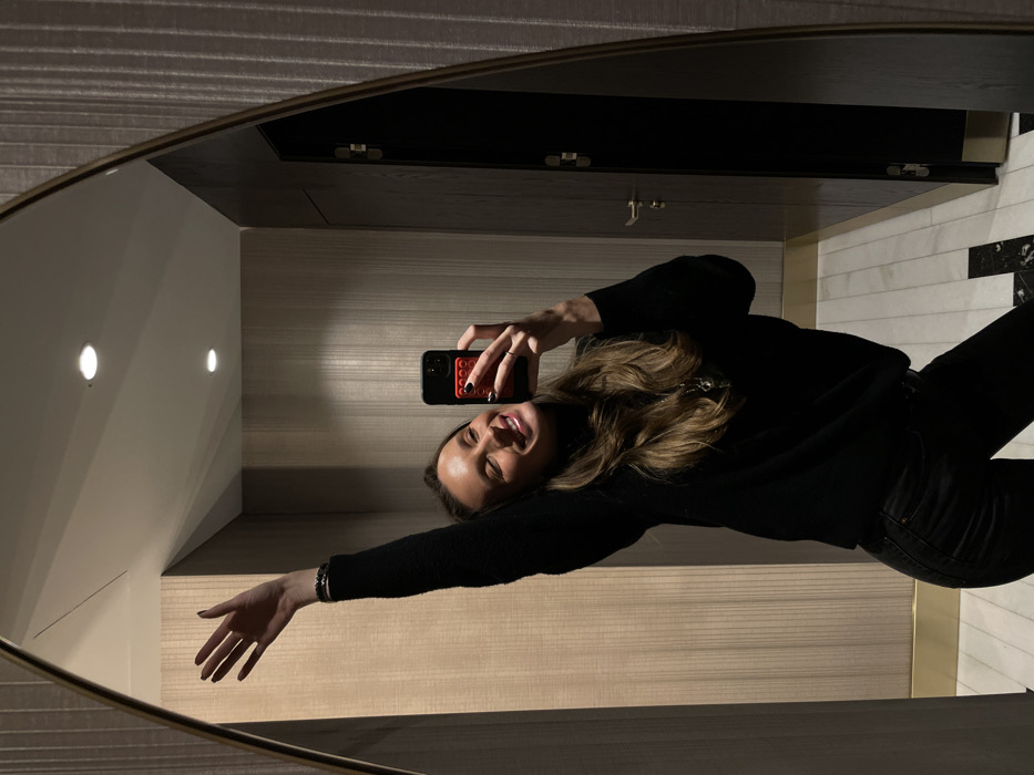 A mirror selfie capturing a woman in a black sweater and jeans. She holds a phone with a red phone case. The background is a modern bathroom with striped walls, a black cabinet, and a marble-tiled section. The lighting is dim. The woman appears to be smiling, with her eyes closed and face angled slightly towards the phone. Her hair is blonde.