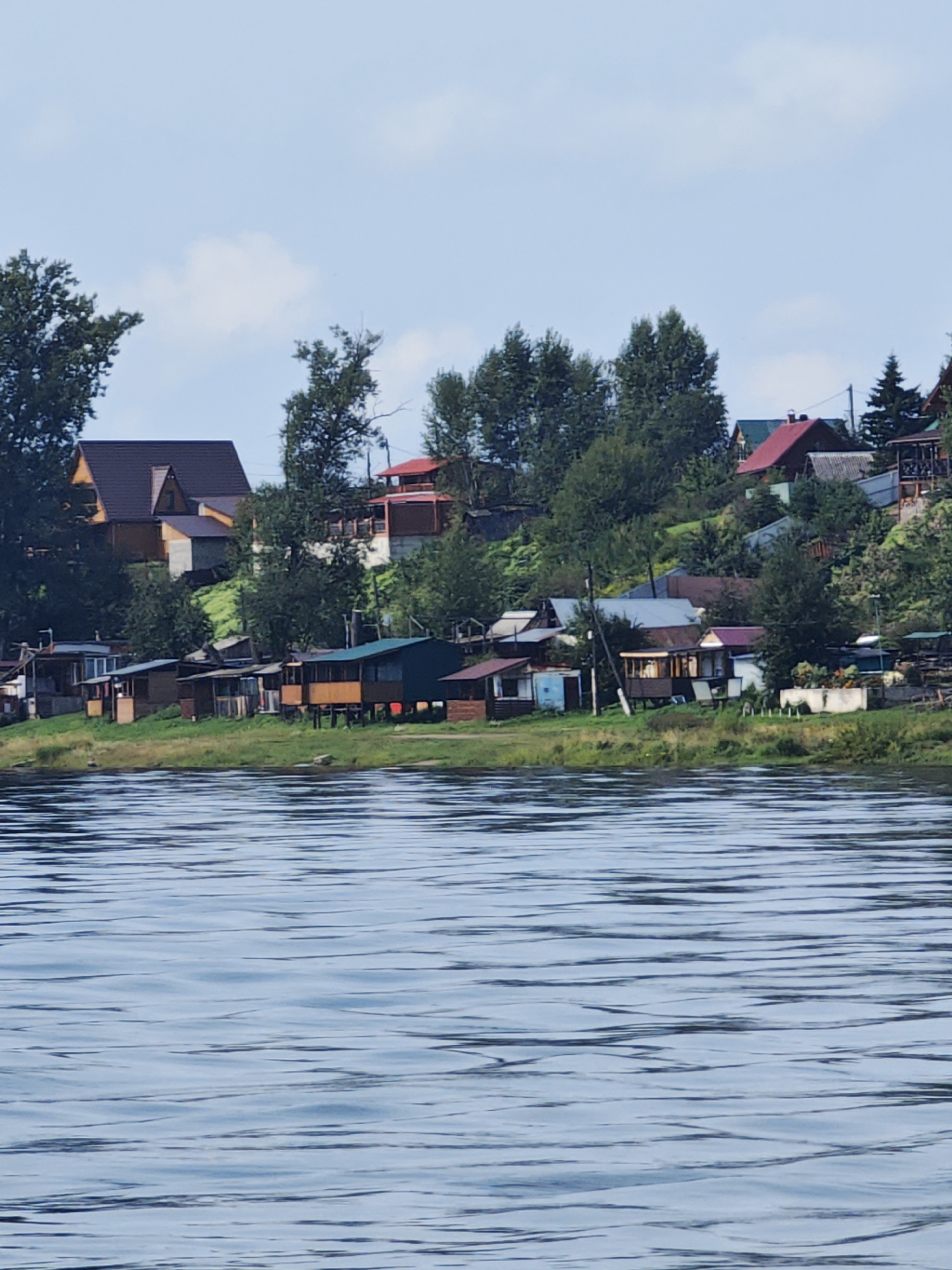 This is a vertical, medium shot of a waterfront village in Kononovo, Russia. The foreground is dominated by rippling blue water. Behind the water, on a grassy embankment, is a cluster of houses and small buildings. The buildings vary in size and color, with some having wooden siding and others with darker roofs. Trees are interspersed among the buildings and on the hillside behind them. The sky above is a pale blue with a few scattered white clouds. The overall impression is a quiet, rural scene.
