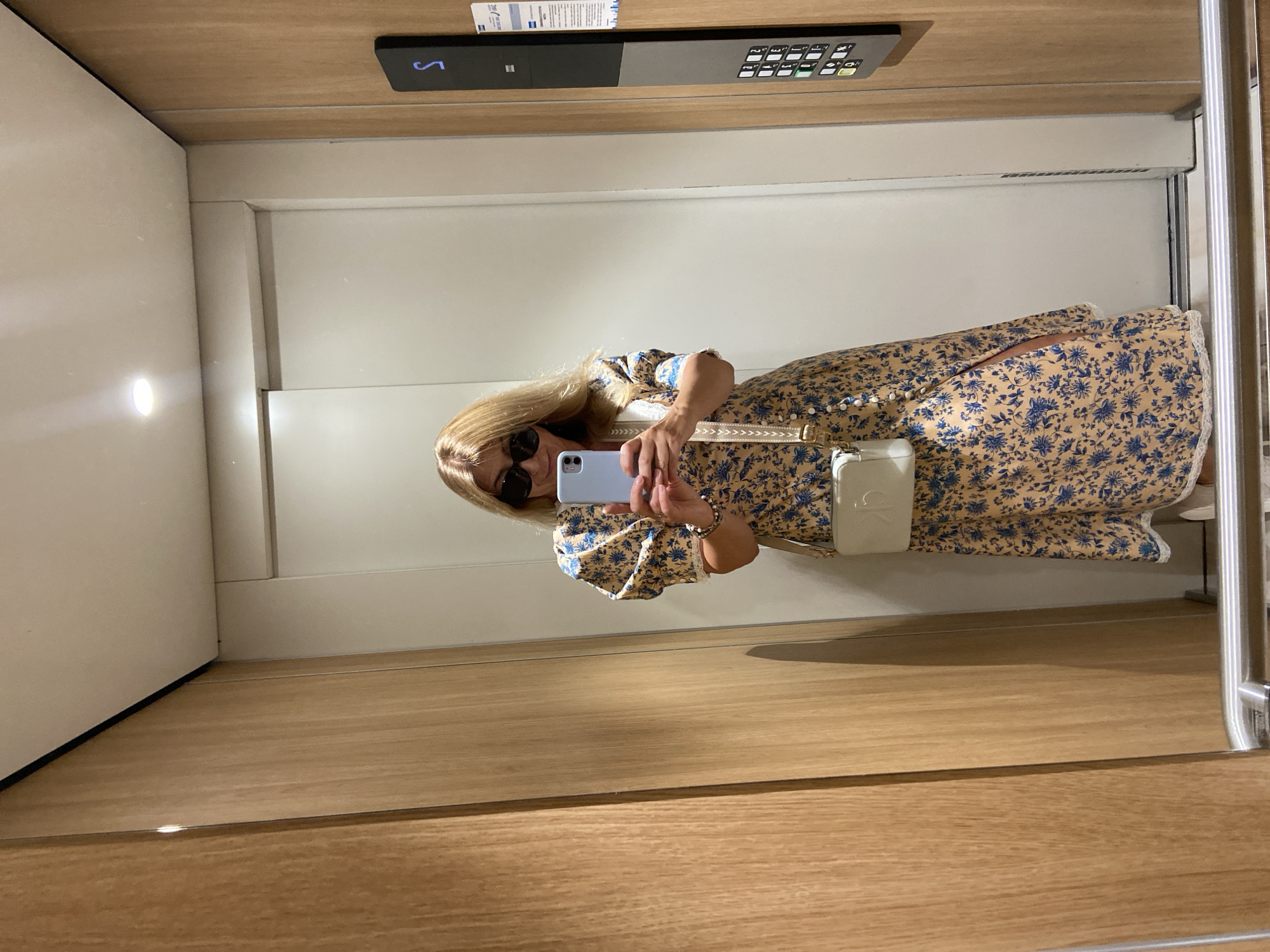 The image shows a woman in an elevator in what could be Budva, Montenegro, based on general architectural styles and available public information. The woman is fair-skinned with long blonde hair and wearing sunglasses. She is wearing a floral-print dress with a slit and carrying a small white purse with a visible brand logo. She is holding a light blue smartphone in her hands, seemingly taking a selfie in the elevator mirror.
The elevator has a modern interior with light wood paneling on the lower portion and light-colored, possibly white, walls above. There is an elevator control panel visible at the top of the frame. The lighting is bright, likely from overhead sources within the elevator cabin.