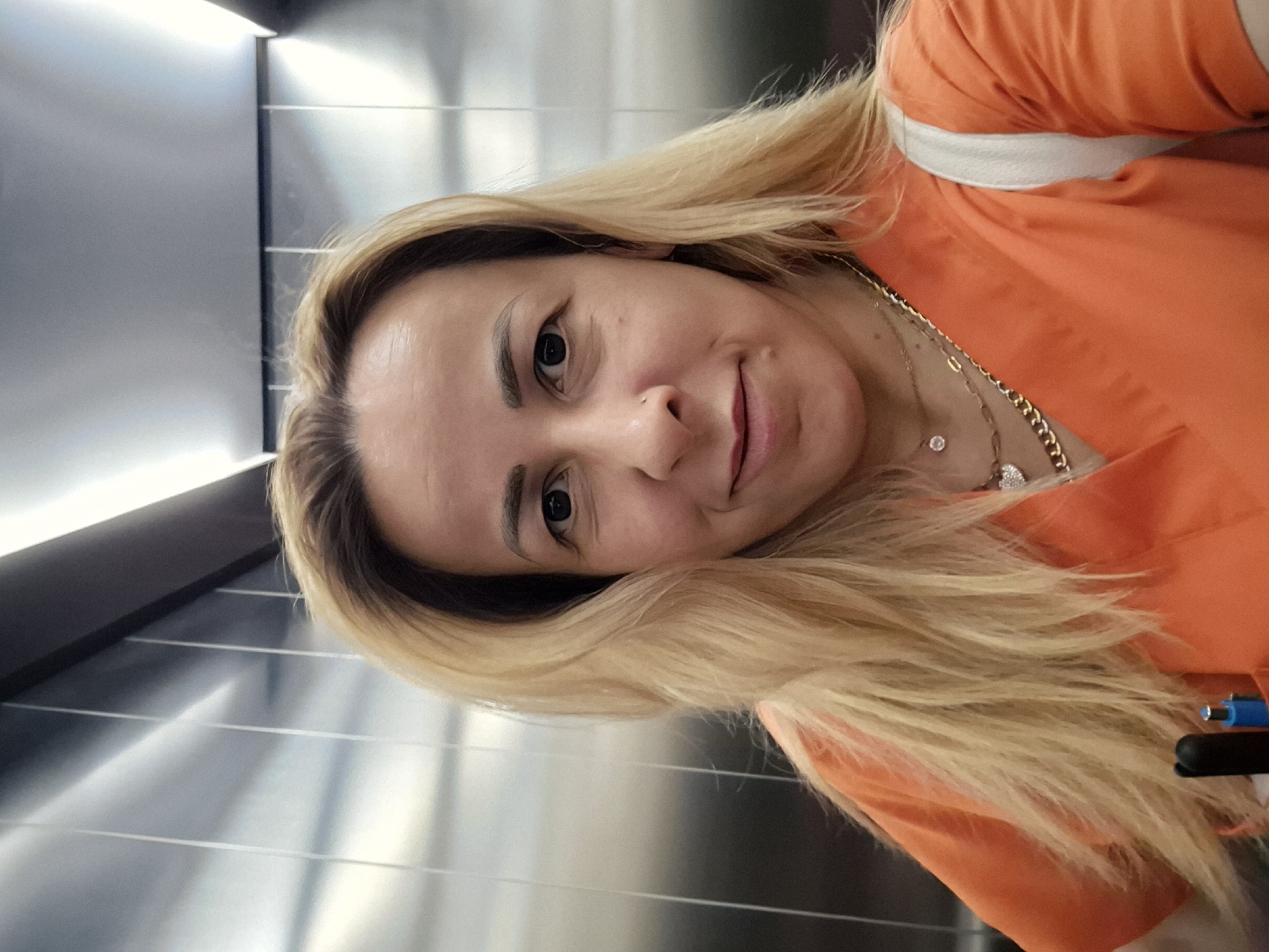The image is a tilted, close-up selfie of a woman with long, flowing blonde hair that has darker roots. She looks directly at the camera with a slight smile and dark eyes. Her face features well-groomed eyebrows and a small nose piercing on her right nostril. She is wearing an orange top, possibly a scrub top or casual shirt, with a white band visible across her shoulder. Several gold-colored necklaces are layered around her neck, including one with a small sparkling pendant and another with a heart-shaped charm.
The background consists of sleek, metallic panels with prominent horizontal lines, suggesting an indoor setting like an elevator, a modern hallway, or a contemporary room. Linear artificial light sources are visible along the top and bottom edges of the metallic walls, creating reflections. On the far right, near her arm, parts of a blue and a black pen-like object are visible. The woman appears calm and poised. No text is visible in the image.