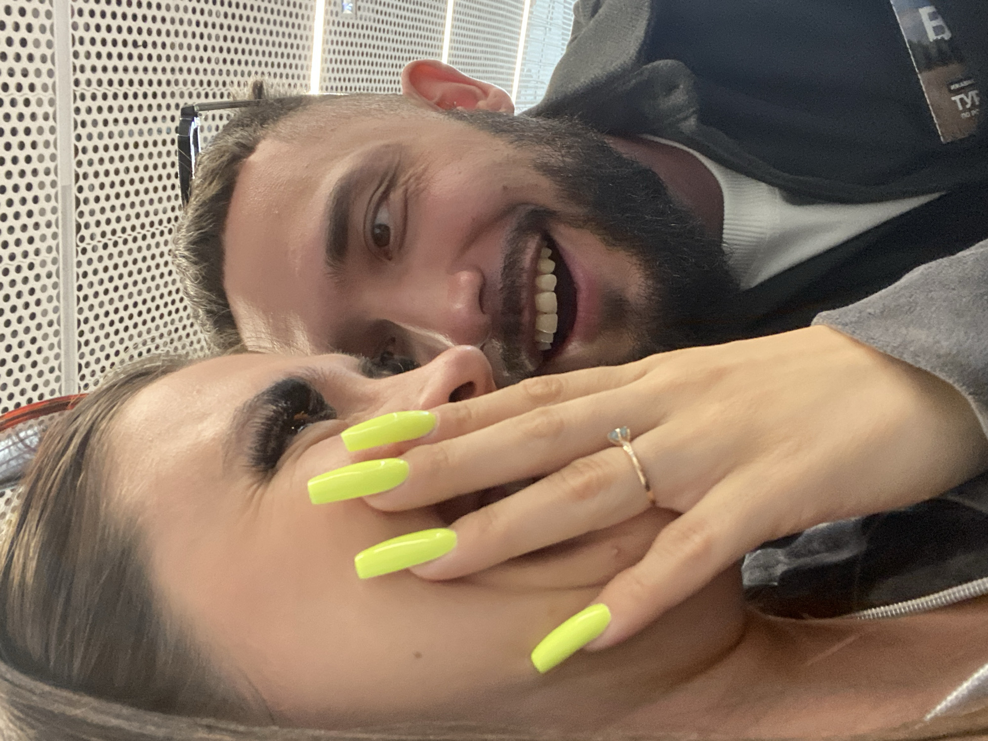 A close-up shot captures two individuals, a woman and a man, sharing a moment. The woman, on the left, has bright neon yellow nails and is holding her hand to her face, revealing a ring on her finger. Her eye is partially visible, adorned with long eyelashes. The man, on the right, has a beard and is looking to the side with a wide, open-mouthed smile. Both appear to be indoors, with a textured, perforated ceiling visible in the background. The scene is likely from the city of Новое Мартьяново, Russia.