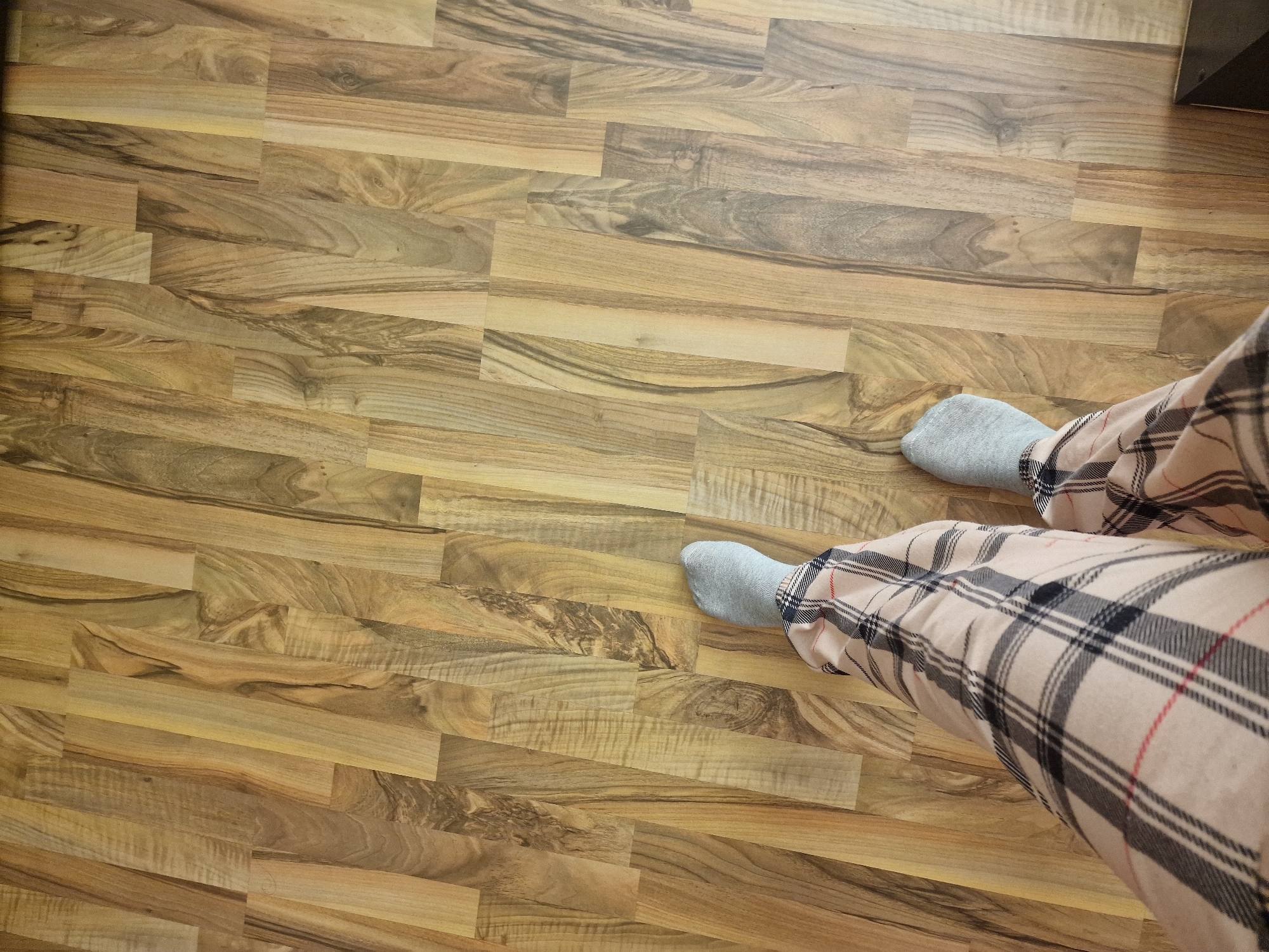 A top-down shot shows a person wearing light gray socks and tan plaid pajama pants. The person is standing on a floor made of light and dark brown wood laminate planks. The location is an interior space, but the city and other identifying details are unknown.