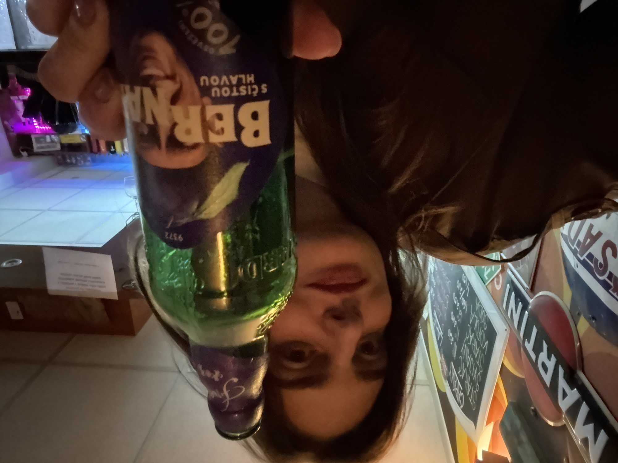 A person is holding a bottle of "Berna S Cistou Hlavou" alcohol. It is green glass with a purple label that reads, in part, "Berna S Cistou Hlavou 100%". The person is in what appears to be a bar setting. In the background, there are signs with the word "Martini" visible, along with other menu boards. The bar area has a light-colored tiled floor and a wooden bar top. Other bar accoutrements are visible in the background. The location is likely in Trnava, Slovakia, given the Czech/Slovakian language on the bottle.