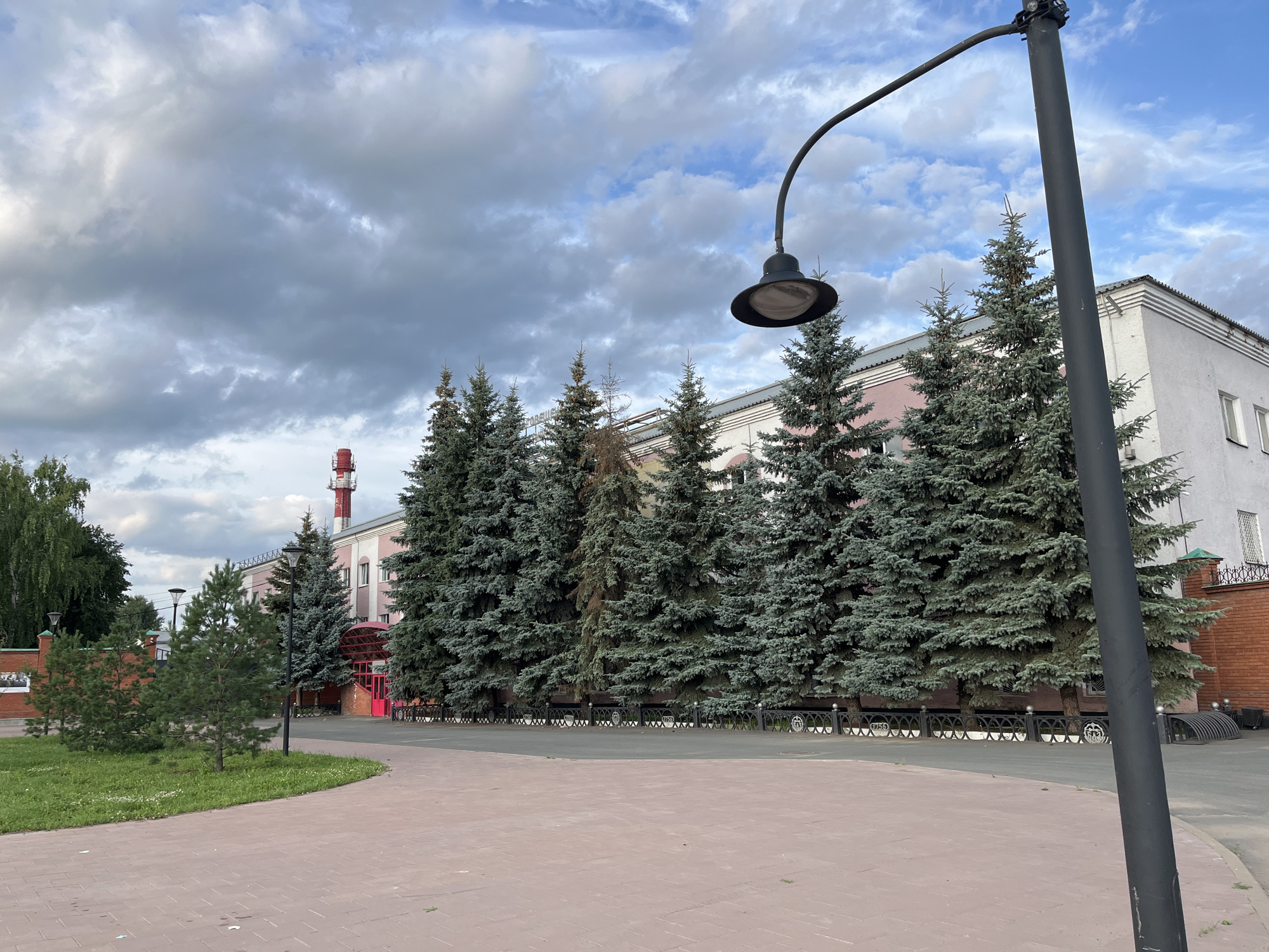 The image shows a street lamp in the foreground, with a paved area in front of it. Behind the paved area is a lawn and then a row of coniferous trees, mostly fir trees. Beyond the trees is a building with a pink and white facade, and a red-roofed chimney visible in the background. A black metal fence runs along the front of the building. The sky is partly cloudy. The scene is consistent with an outdoor area in the city of Blagoveshchensk, Russia, possibly a public space or the grounds of an industrial facility. There are no people visible in the image.