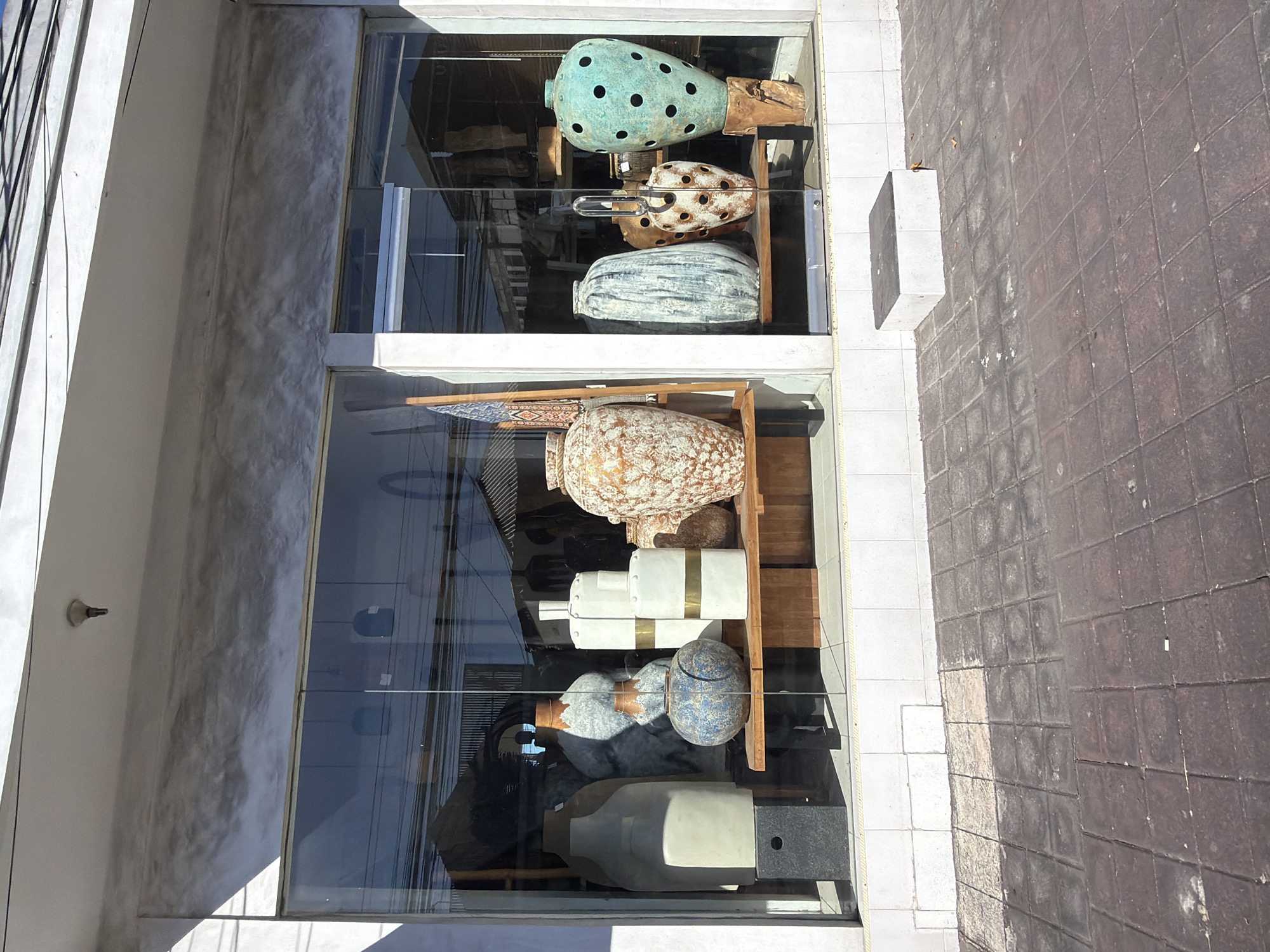 The image depicts a street-level view of a storefront in North Kuta, Indonesia. The storefront features large glass windows displaying various ceramic vases and objects arranged on shelves. The objects vary in color, size, and design, including speckled, dotted, and banded patterns. The windows reflect the exterior environment, creating visual layers and distortions. The exterior of the building exhibits a textured facade. A paved sidewalk runs adjacent to the building, with tiles varying in color and pattern. No people are visible in the immediate scene.