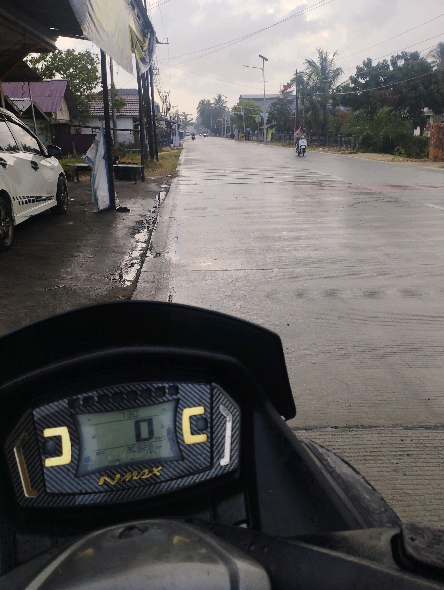 The image is taken from the perspective of someone riding a motorcycle on a paved road in Kuala Samboja, Indonesia. In the foreground, the motorcycle's digital dashboard is visible, displaying a speed of 0 km/h and an odometer reading of 30928 km. The dashboard has a carbon fiber-like texture with yellow accents and the word "Nmax" in gold lettering.
The road ahead is wet and appears to be a two-lane street with faint white road markings. The sky is overcast, suggesting a cloudy or possibly rainy day. On the left side of the road, there is a white car parked, and a small building with a purple roof. A tall utility pole stands next to the road, with wires running overhead. Further down the road, a person is riding a white scooter. On the right side of the road, there are trees, a concrete barrier, and a small brick structure. The overall scene depicts a somewhat rural or suburban street in a tropical climate.