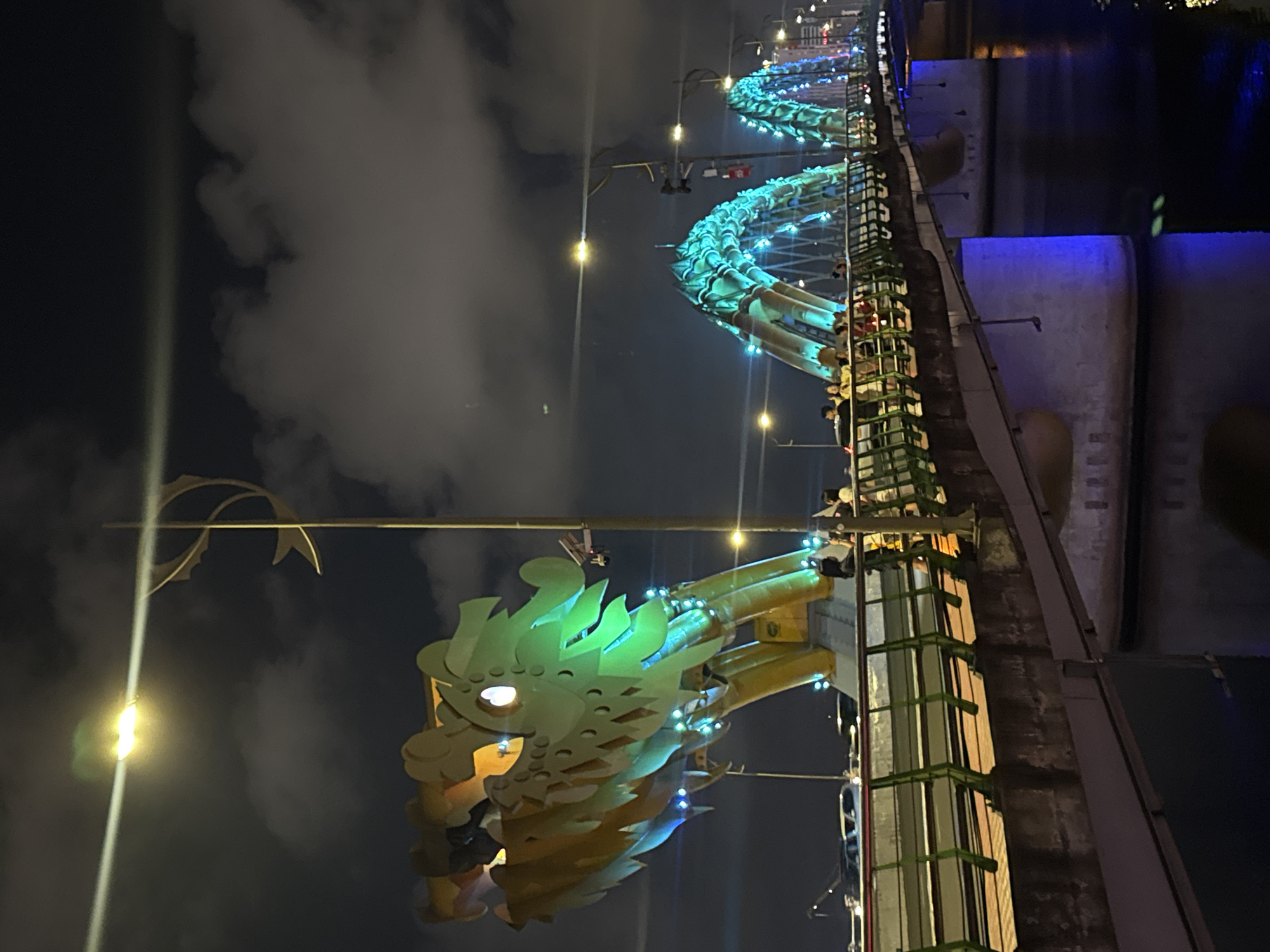 The image shows the Dragon Bridge in Đà Nẵng, Vietnam at night. The illuminated dragon head at the bridge's eastern end is prominent. The bridge's arching structure extends into the distance, also illuminated, above a body of water. There is a walkway along the side of the bridge, where several people are visible. Streetlights are interspersed along the bridge's length, adding to the scene's overall brightness. The sky is dark, and clouds are visible.