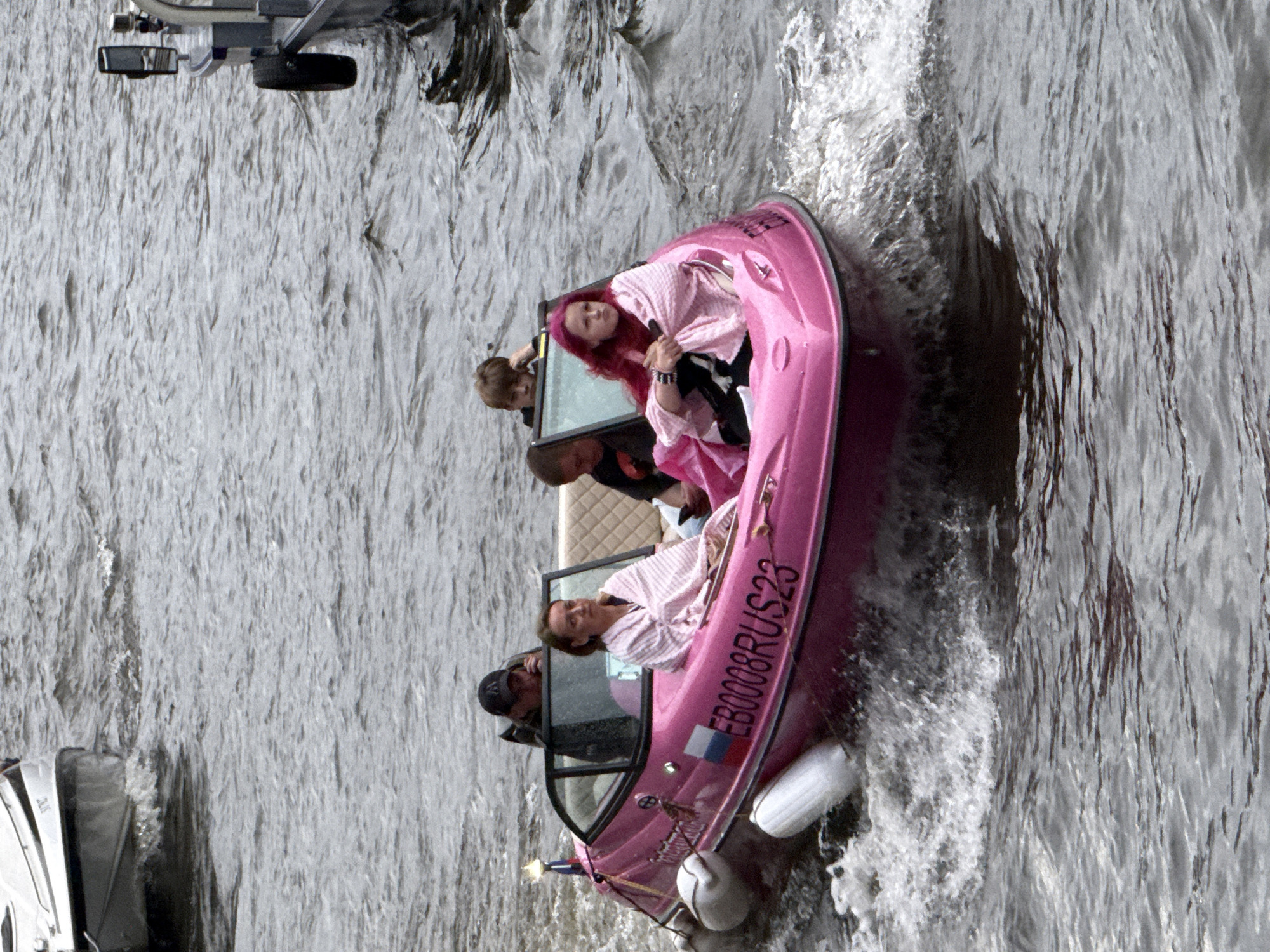 A bright pink motorboat navigates a body of water, likely the Neva River in Saint Petersburg, Russia, given the presence of Russian flag markings on the boat. The boat contains four individuals. In the front, a young person with bright pink hair is wrapped in a pink and white striped blanket. To their right, a woman with brown hair, also wrapped in a similar blanket, sits attentively. Behind them, a man in a black baseball cap appears to be speaking on his phone, and to his right, a young boy with brown hair is leaning forward. The boat is creating a significant wake as it moves, and other boats can be seen in the background. The water is choppy, indicating a moderate level of current or wind. The overall scene depicts a recreational outing on the water in an urban setting.