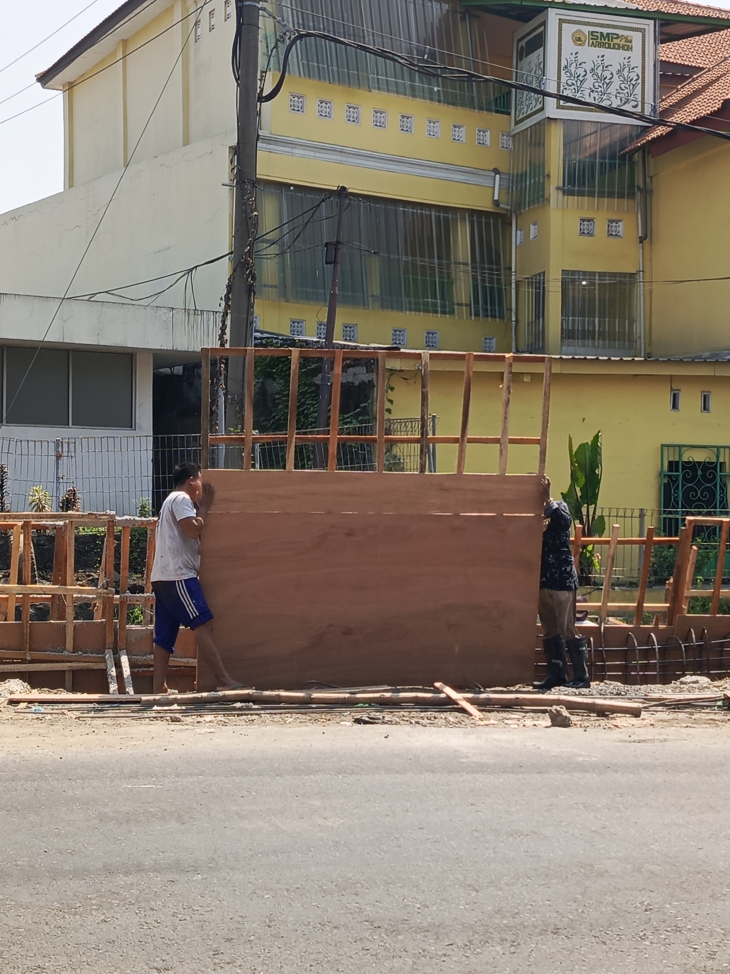 In Wedi, Indonesia, two men are engaged in construction work on a street. One man, wearing a white t-shirt and blue shorts, is pushing a large wooden board. The second man, wearing a dark patterned shirt and dark pants tucked into black boots, is positioned behind the wooden board, appearing to assist. They are working with a wooden frame structure that supports the large board. In the background, a yellow building with a prominent sign reading "SMP N 1 ARROUDHOH" is visible, along with power lines and vegetation. The scene suggests an outdoor construction project taking place adjacent to a road.
