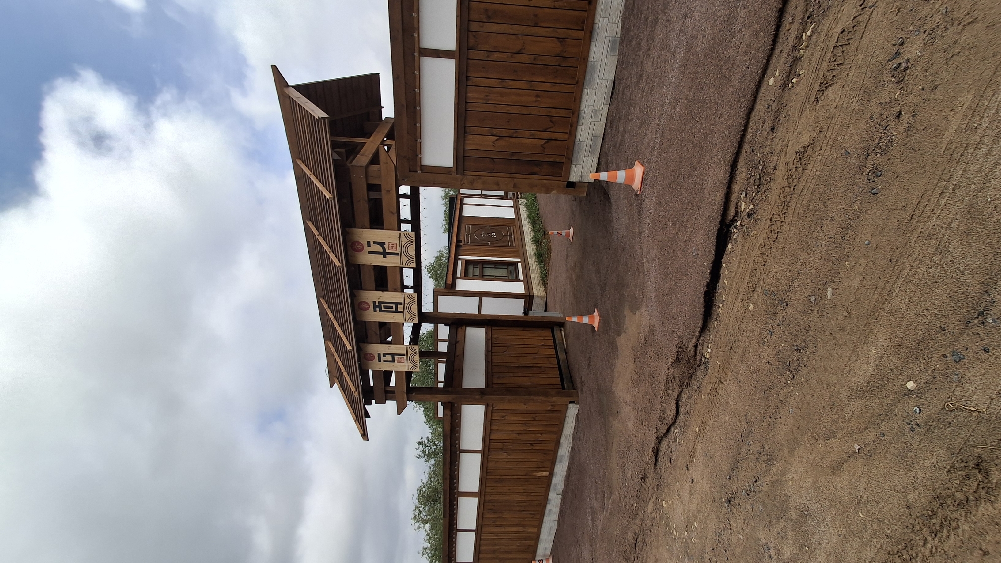 The image depicts a structure with Asian architectural influences, likely a pavilion or gate, situated in what appears to be a park or recreation area. The structure is primarily constructed of wood, with white panels integrated into the design. It has a multi-tiered roof decorated with dark colored ornamentation that suggests oriental or traditional design motifs. Two orange traffic cones stand near the structure. The ground is covered with gravel and dirt. The sky is overcast with light clouds. The location is Kempelevo, Russia.