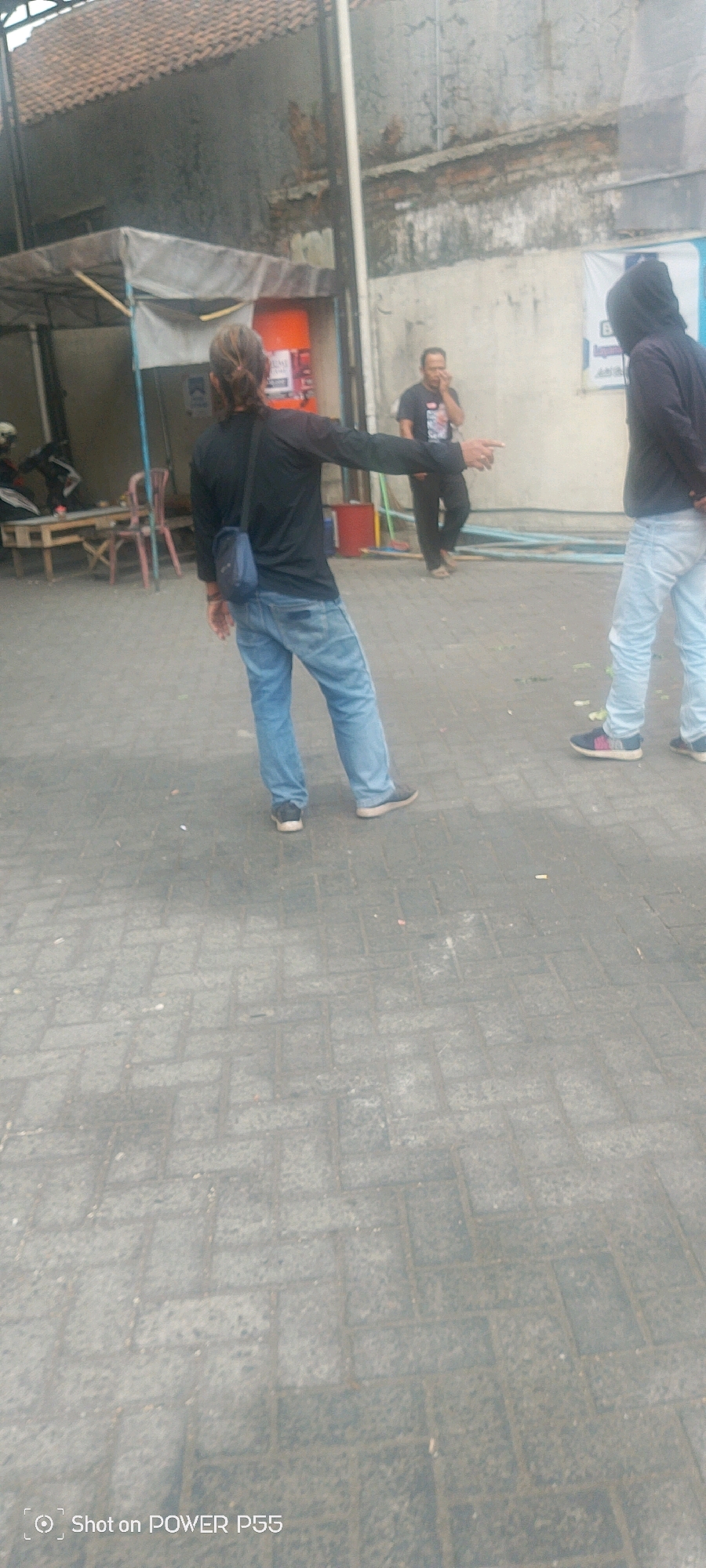 In this full shot, two men are visible from the waist down on a paved walkway. The man on the left is wearing a black long-sleeved shirt, blue jeans, and dark sneakers. He has a blue crossbody bag and is pointing to the right. The man on the right is wearing a dark hooded jacket and light blue jeans, with dark sneakers. He is standing with his hands at his side. The environment suggests an outdoor urban setting with a tiled pavement and a building in the background.