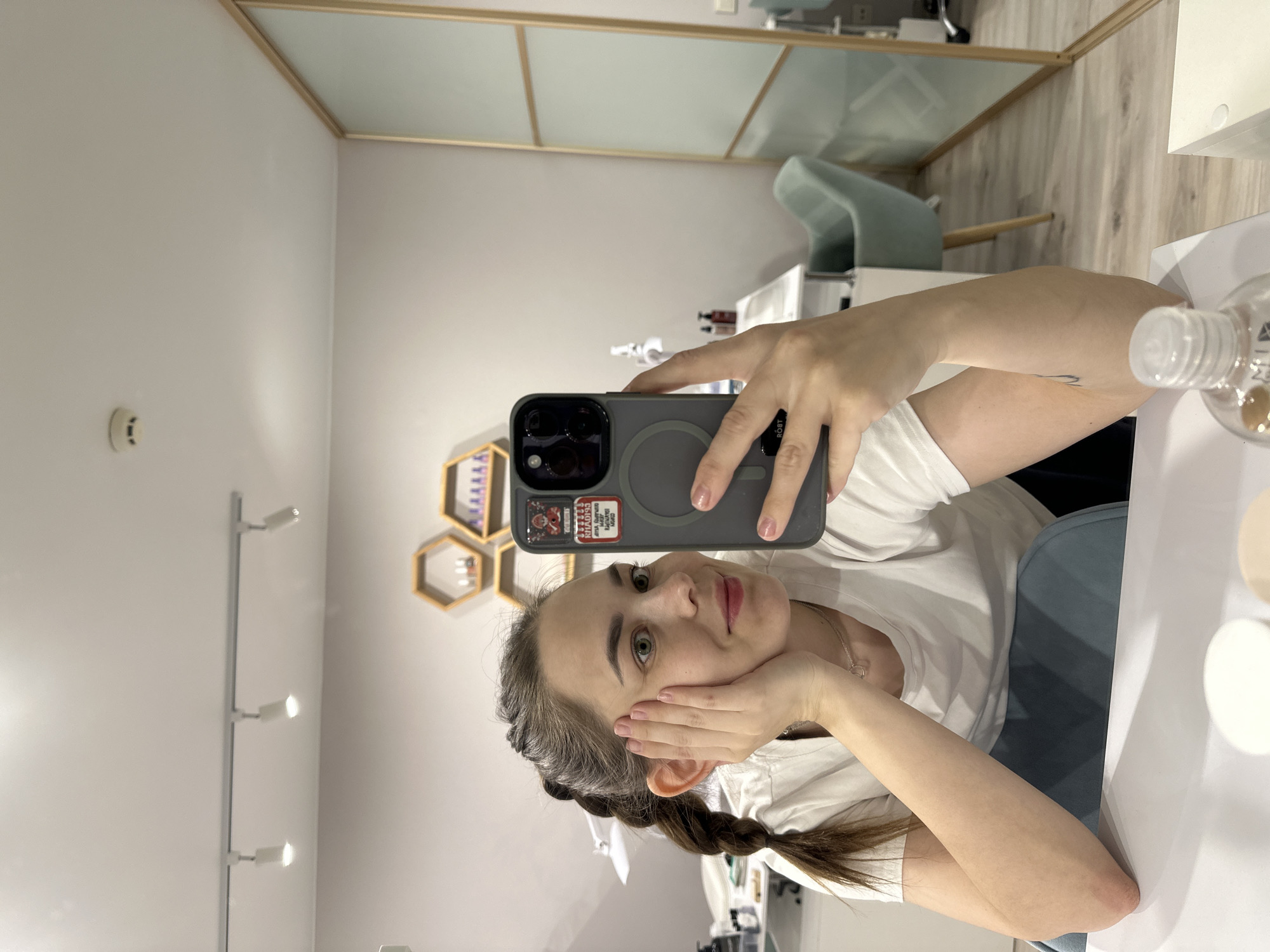 A woman is taking a selfie with a gray iPhone in what appears to be a cosmetic salon in Perm, Russia. She is wearing a white T-shirt and her hair is styled in braids. Her left hand is touching her face. The background shows a white table and a wall with a display of hexagonal shelving. A light fixture is visible on the left wall. A divider wall with glass panels and a green chair are in the background. Several small bottles and jars are on the table to the right.