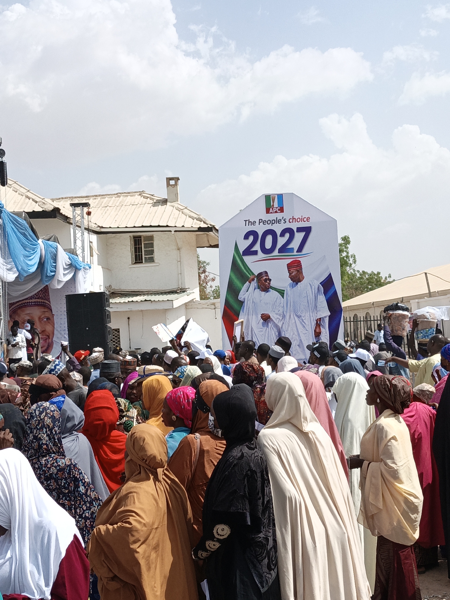 This image depicts a large outdoor gathering, likely a political rally, in Giginyu, Nigeria. A significant crowd of people, primarily adults, occupies the foreground and midground, facing towards a stage area and prominent banners. Many attendees are dressed in traditional attire, including various head coverings such as hijabs, turbans, and caps, in a spectrum of colors and patterns.
The central feature is a large white political banner or billboard. It prominently displays the "APC" logo, the slogan "The People's choice," and the year "2027" in bold lettering. The banner features an illustration of two men in white traditional garments and red caps, standing side-by-side, against a backdrop incorporating green and white, reminiscent of the Nigerian flag.
To the left, a stage is adorned with flowing blue and white fabric. A smaller poster showing the face of another man is visible on this side, alongside large black speakers mounted on a truss structure, indicating sound amplification for the event. In the background, a white building with a light-colored tiled roof and a chimney stands. Another tent structure is partially visible on the far right.
The scene unfolds during the daytime under a bright sky dotted with scattered white clouds, suggesting clear weather. The crowd appears engaged, observing the proceedings. Some individuals are seen carrying items on their heads. The overall context points to a political event or campaign focusing on future elections in 2027.