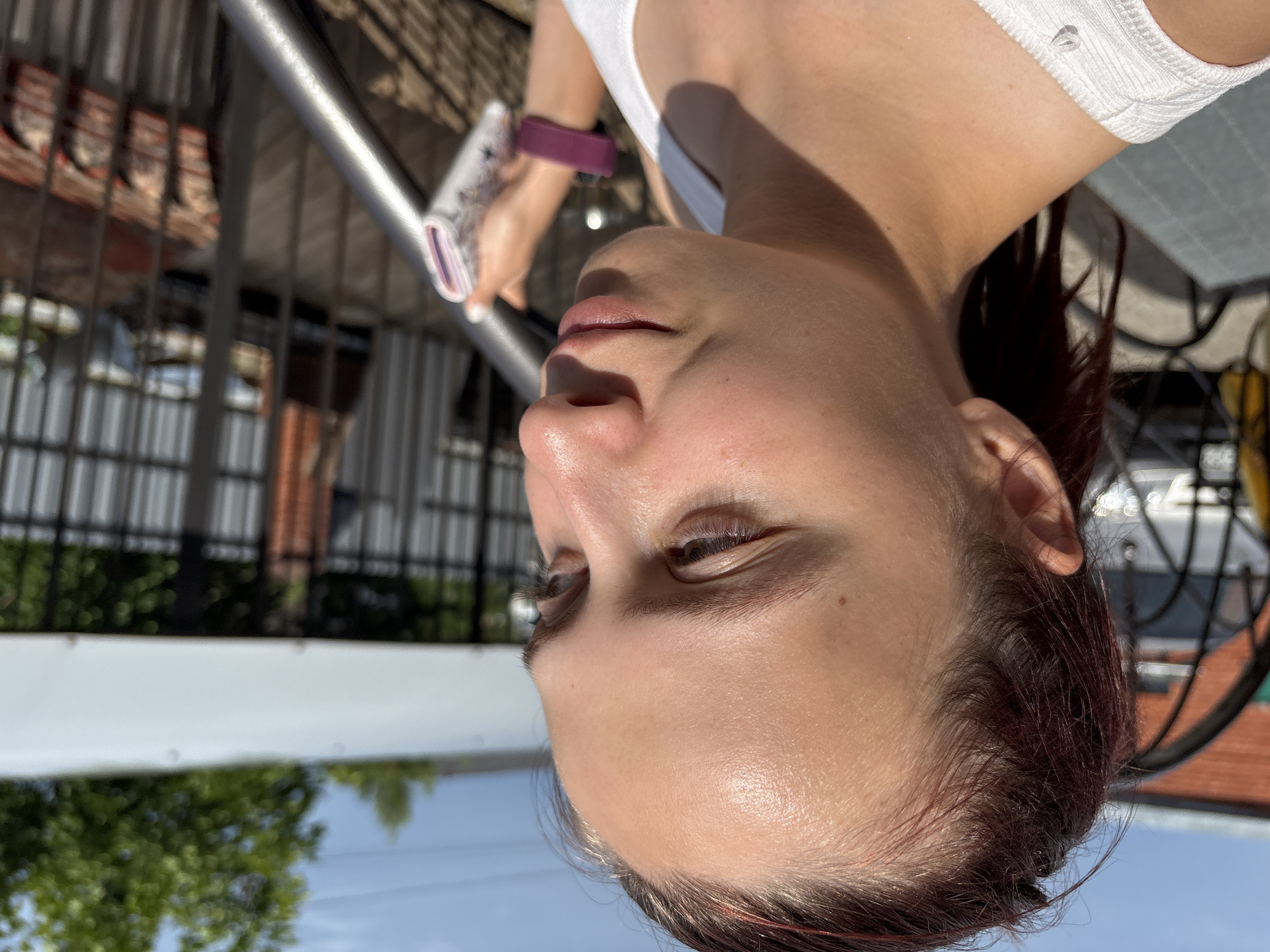 A young woman with dark hair pulled back is shown in profile, looking to her right. She is wearing a white tank top and a purple fitness tracker on her left wrist. She is holding a small, white, rectangular object with a colorful design in her right hand. The background suggests an outdoor setting with a metal fence, a white roof structure, trees, and a brick building visible. The lighting indicates it is daytime. The image appears to be a candid photograph taken outdoors, possibly in a residential or communal area. The location "Прикамский, Russia" is noted, but the image itself does not contain any specific visual cues to confirm this location.