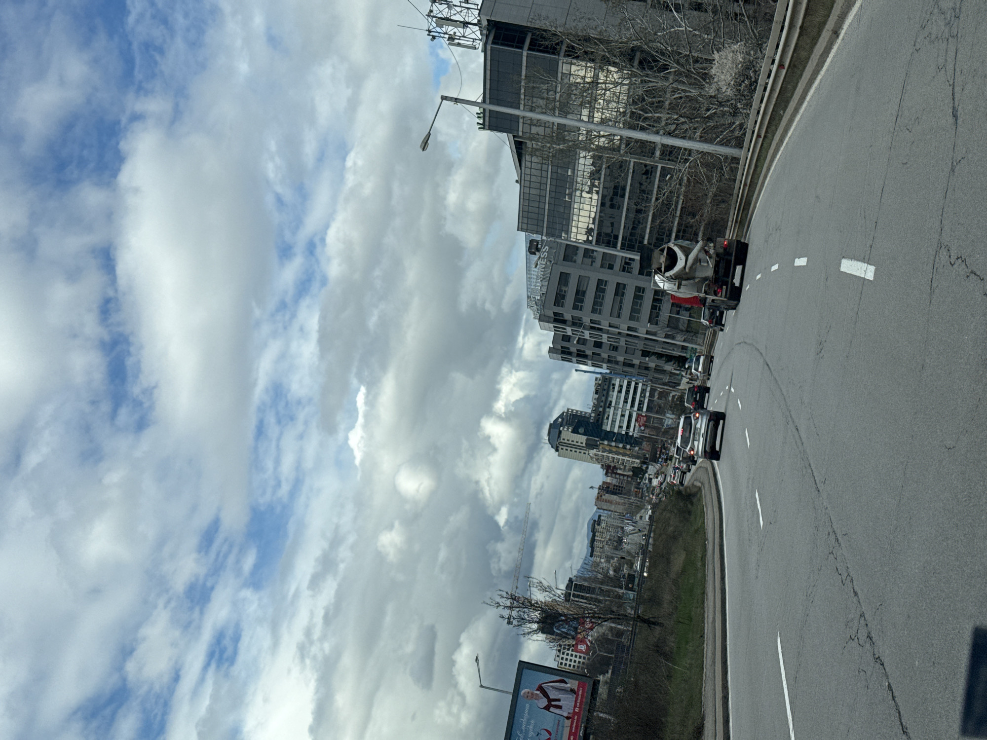 The image depicts a street scene in Sofia, Bulgaria. A highway curves slightly to the right, with several cars and a concrete mixer truck visible on the road. The road is marked with dashed white lines. To the left of the highway are various buildings, including modern office blocks and residential apartments. In the distance, there are mountains and a cloudy sky. A billboard is visible on the left side of the road. The sky is filled with white and gray clouds. The overall perspective suggests the photo was taken from a moving vehicle.