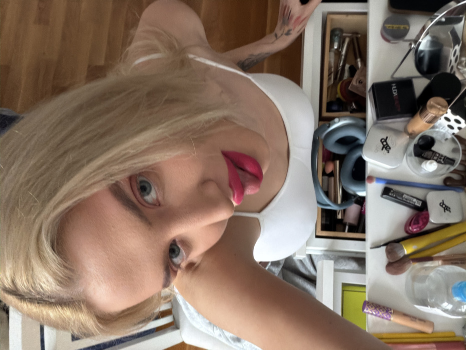 An overhead shot depicts a fair-skinned individual with long, straight blonde hair, light blue eyes, and prominent pink lipstick. The individual wears a white spaghetti-strap top and appears to be leaning over a white multi-compartment storage unit. The tongue is slightly extended. A tattoo, possibly floral, is visible on the upper right arm/shoulder. The individual's left arm is partially visible, resting on an unseen surface to the left. Below the individual, the storage unit's open compartments reveal various makeup items, including several brushes with yellow and natural wood handles, a Huda Beauty compact, other cosmetic containers, a small round mirror, and a pair of blue/grey over-ear headphones. The background consists of a light-colored wooden surface to the right and a white and striped object, possibly a chair back or headboard, to the left.