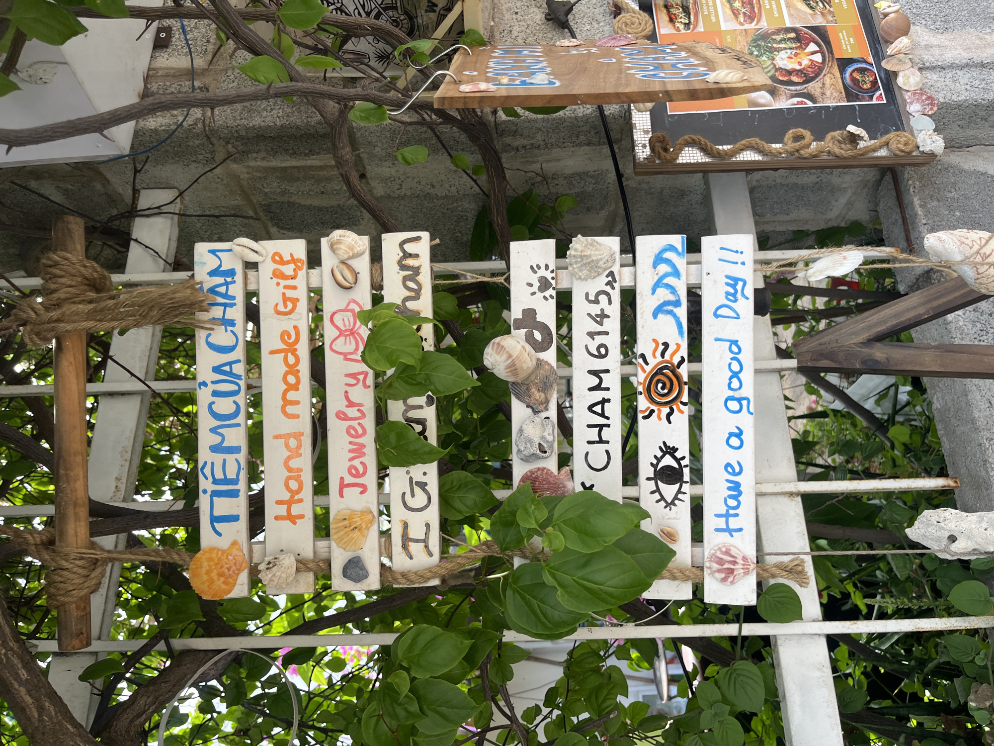 A white wooden sign with multiple slats is displayed outdoors, suspended by rope from a tree branch. Each slat contains text or images. The top slat reads "TIÊM CỦA CHAM" in blue lettering. Below that, "Hand made Gift" is written in orange. The next slat has "Jewelry" in pink, accompanied by a drawing of a pink bow. Another slat displays "I G : CHAM" in dark brown. The following slat reads "CHAM 6145" in black. Above this, there are depictions of an eye and a sun with waves. The bottom slat says "Have a good Day!!" in blue. The sign is decorated with various seashells and stones attached to some of the slats. Lush green foliage surrounds the sign, suggesting an outdoor, possibly tropical, setting. A wooden menu board with pictures of food items is visible to the right of the sign. The overall scene appears to be in Mũi Né, Vietnam, likely a shop or restaurant entrance. There are no people visible in the image.