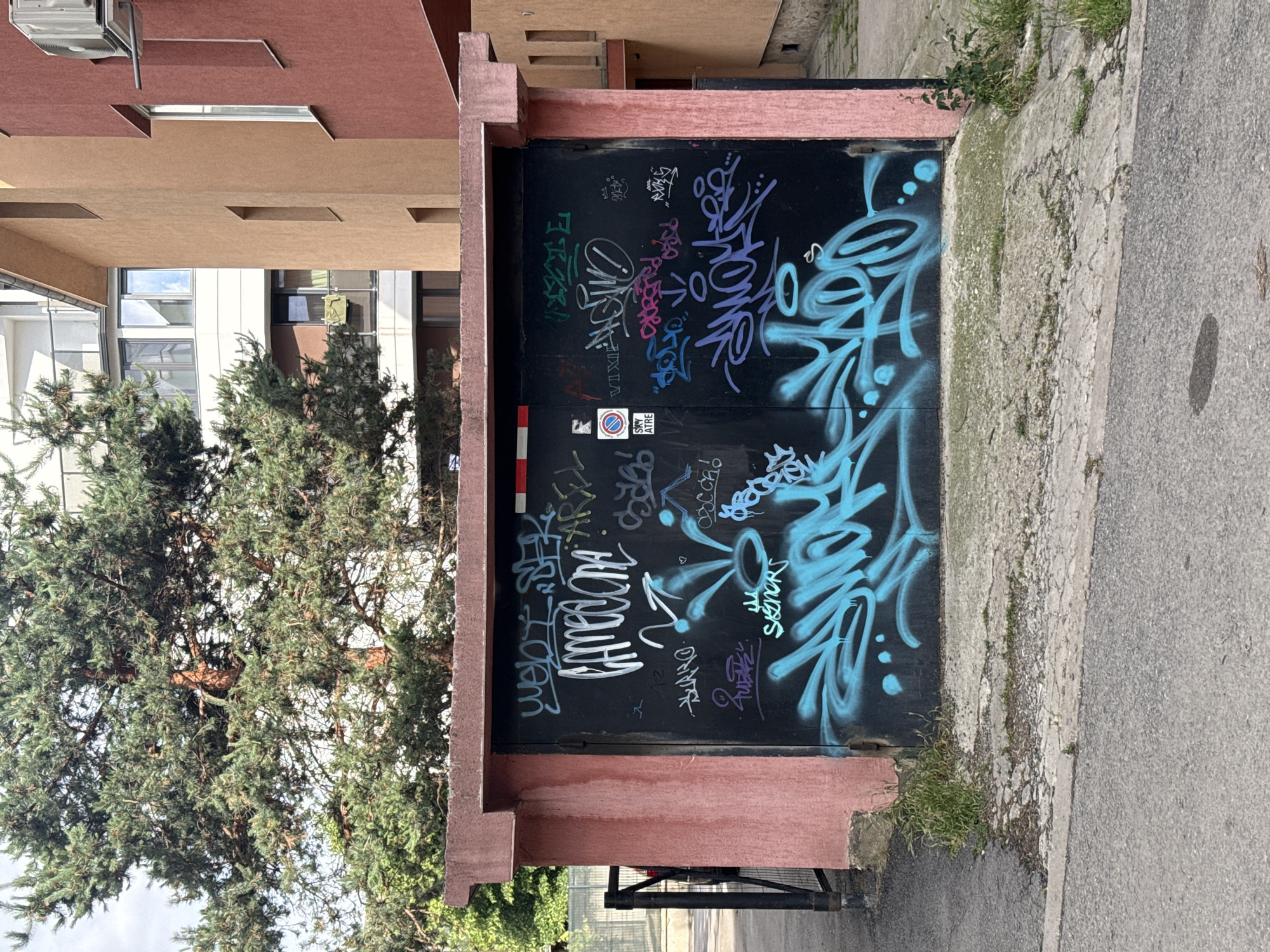 The image shows a grey asphalt road in front of a weathered pink concrete structure with a black metal garage door. The garage door is heavily graffitied with various tags and designs in multiple colors, including white, green, purple, and bright blue. A prominent white tag reads "GAMEONE" in large, bold letters. In the upper left corner of the garage door, there is a red and white striped sticker. To the left of the structure, a black metal fence is visible. Behind the pink structure, a large pine tree with dense green needles is present. Further back, modern multi-story buildings with balconies and windows can be seen, one of which has an air conditioning unit mounted on its exterior. The overall scene appears to be an outdoor urban environment.