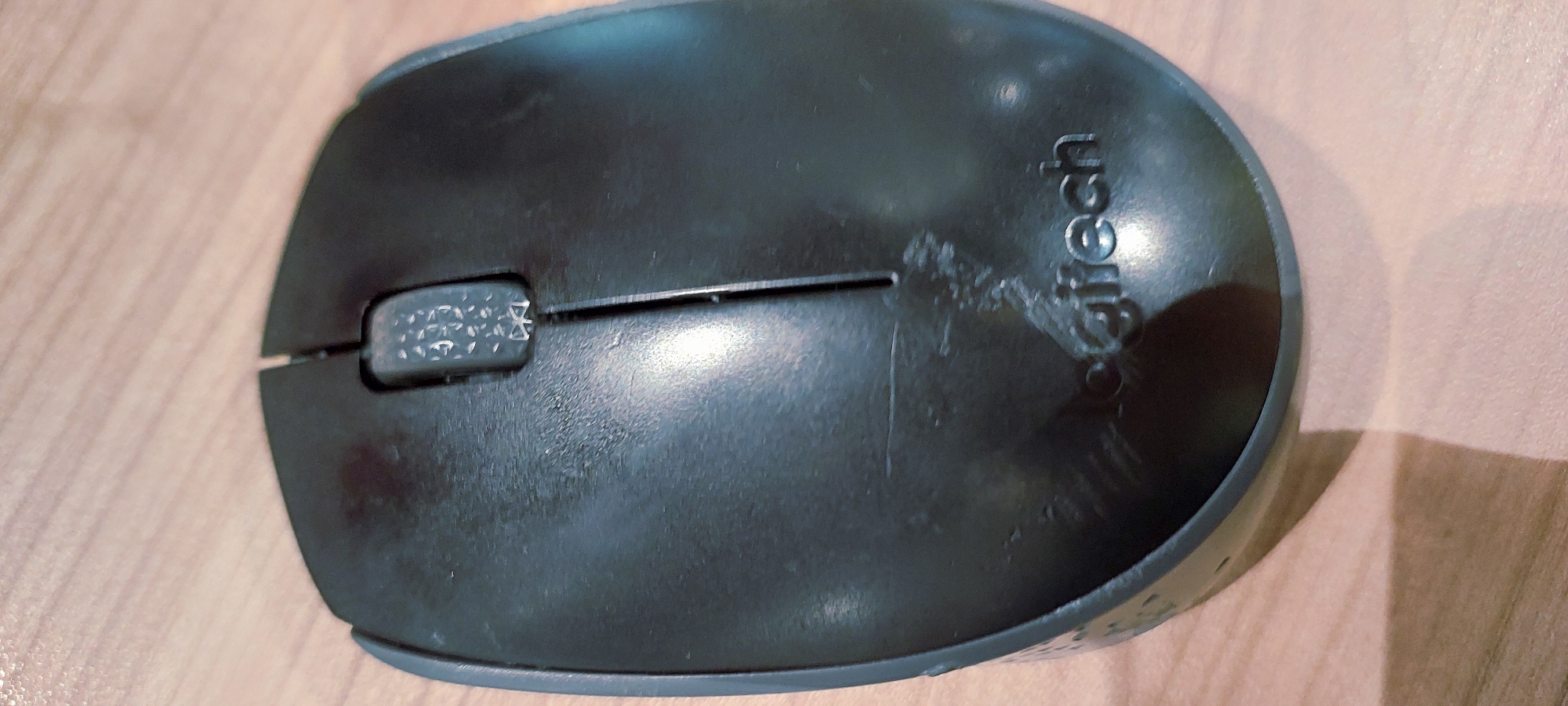 The media file shows a close-up, overhead shot of a black Logitech wireless mouse resting on a wooden surface. The mouse is positioned diagonally from the bottom left to the top right of the frame. The scroll wheel is visible on the left side of the mouse, with a textured pattern. The "Logitech" logo is embossed on the right side of the mouse, though it appears somewhat worn or obscured by smudges. The wooden surface beneath the mouse has a light brown, grained texture. The lighting suggests an indoor setting, possibly with overhead artificial light creating a shadow on the right side of the image, extending towards the bottom right. There are some scuff marks and smudges visible on the surface of the mouse, indicating it is a used item. No people are visible in the image. The scene is a simple still life of an electronic device.