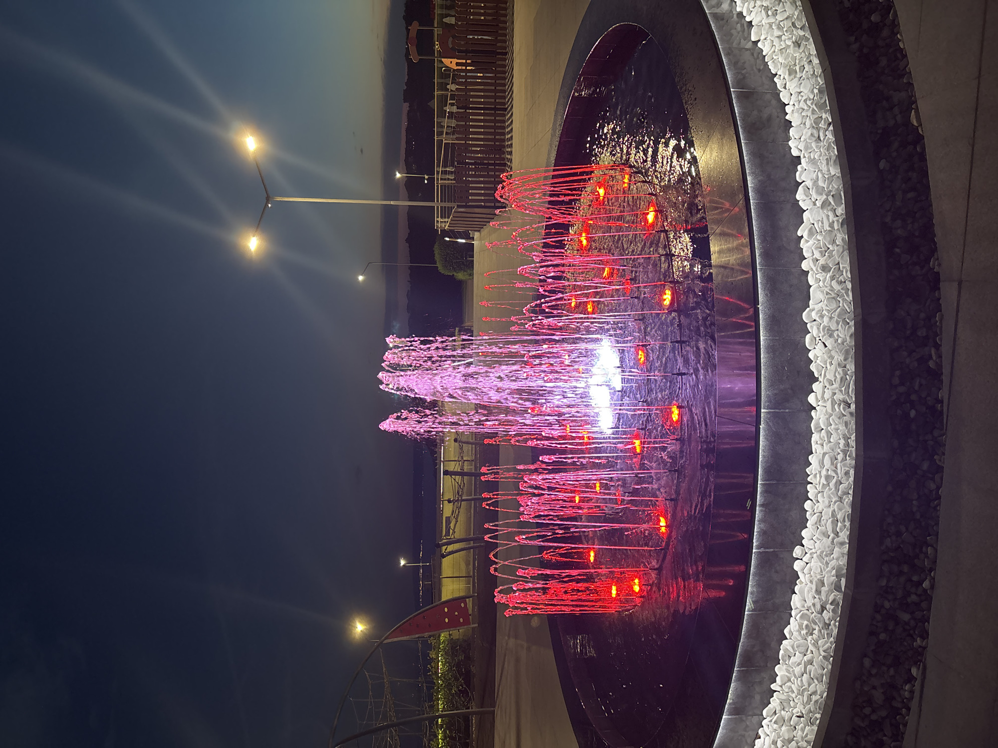 The image depicts a public fountain illuminated with red and pink lights at dusk in the city of Topola, Bulgaria. The fountain is circular with water jets creating arches of light. The surrounding area features white decorative stones, a paved walkway, and a low wooden fence. In the background, streetlights are visible against a twilight sky, along with elements of a playground structure. The scene appears to be an outdoor public space, likely a park or recreational area.