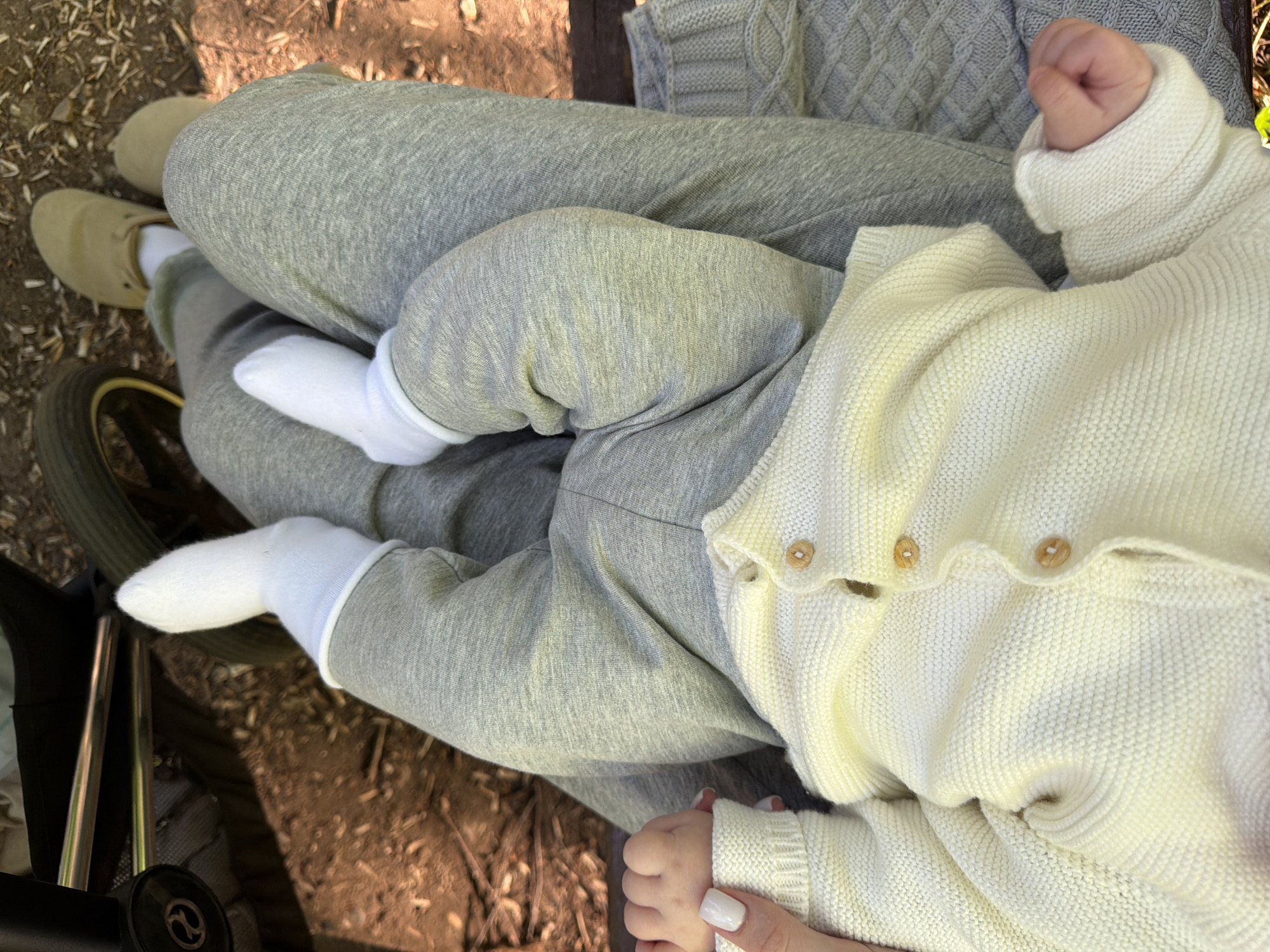 A baby is lying on a person's lap. The baby is wearing a cream-colored sweater and white socks. The person is wearing gray sweatpants and beige shoes. A gray sweater and a stroller are visible in the background. The ground is covered in dirt and leaves. The image was taken outdoors, possibly in a park setting. Location is not possible to ascertain from given image.