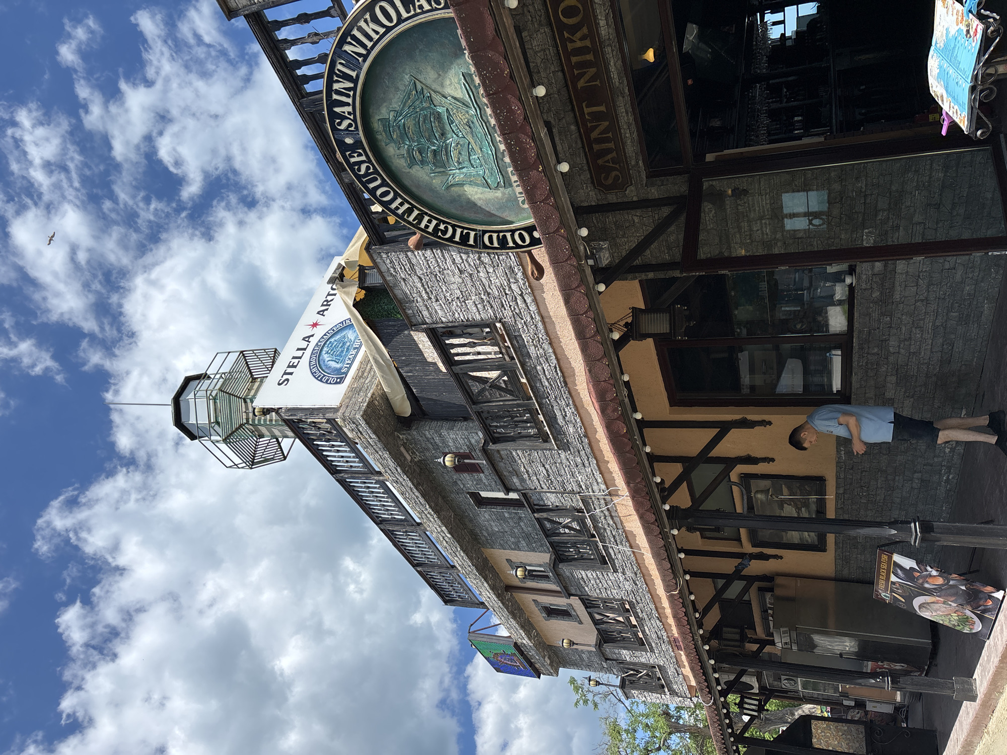 The image captures the exterior of the "Old Lighthouse Saint Nikolao" restaurant, located in Varna, Bulgaria, under a partly cloudy sky. The building features a lighthouse-like structure on top, with multiple levels and balconies adorned with dark wooden railings. The facade is constructed with stone-like grey material and accented with peach-colored elements. A large circular sign with a ship motif and the text "OLD LIGHTHOUSE SAINT NIKOLAUS" is prominently displayed. Below the upper levels, a covered walkway with outdoor seating is visible, featuring a sign advertising "FRESH BLACK SEA FISH." A man in a blue t-shirt and dark shorts stands on the sidewalk, looking at his phone. Various restaurant equipment, such as refrigerators and menus, are arranged outside. The scene suggests a daytime setting, possibly during tourist season, with a maritime theme evident in the restaurant's design and signage.
