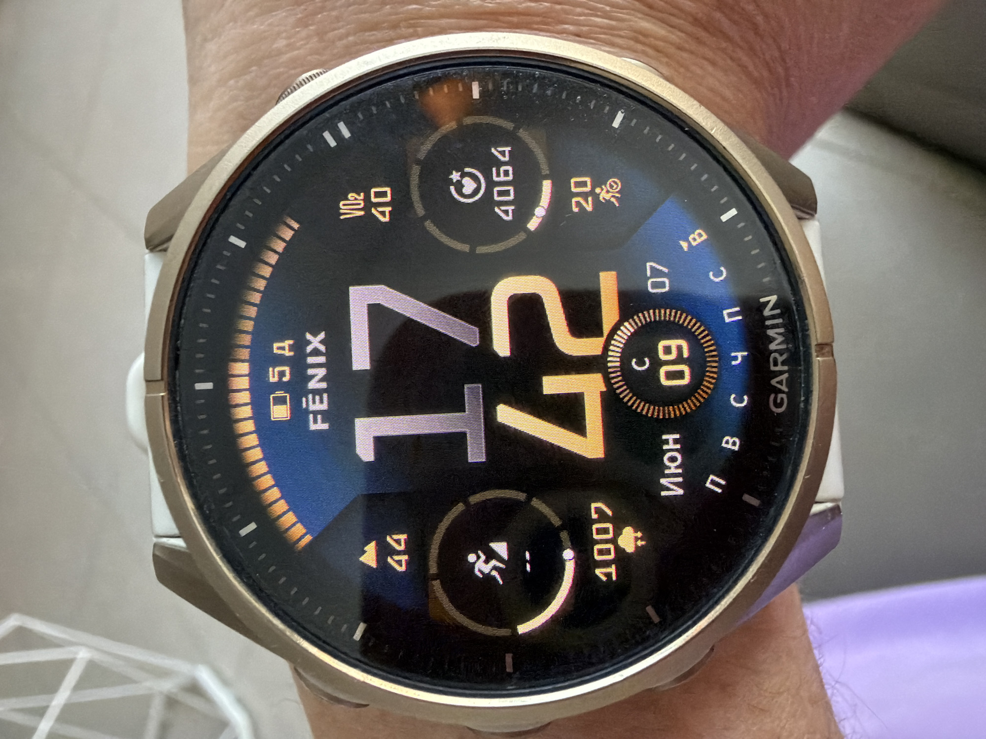 A close-up image depicts a gold-toned Garmin Fēnix smartwatch with a white silicone strap, worn on a person's left wrist. The watch face is digital, black with various data points displayed in grey, orange, and white text.
The time is prominently displayed as 17:42 (5:42 PM), with "17" in large grey numerals and "42" in large orange numerals. Above the time, "FĒNIX" is written in white, below a battery icon showing "5A" (likely indicating battery level or days remaining). A semicircular arc with orange segments is present at the top of the watch face.
Various activity and health metrics are arranged around the main time display:
- In the upper left quadrant, an upward-pointing orange arrow with the number "44" (potentially indicating altitude or temperature) is visible.
- Below that, a running person icon within a circular progress bar, showing "1007" (likely daily activity points or floors climbed) and a cloud with rain icon beneath it. The progress bar is partially filled.
- In the upper right quadrant, "VO2 40" indicates a VO2 Max score of 40.
- Below that, a heart icon with a refresh arrow within a circular progress bar, showing a numerical value of "4064" (likely daily steps or an activity metric). A progress bar below this shows two orange segments filled.
- In the lower right quadrant, a bicycle icon with "20" below it (likely cycling activity time or distance).
The date "Июн 09 07" is displayed in the lower center, with "Июн" being Russian for June, "09" for the day, and "07" an additional numerical value (possibly temperature or year indicator). Below this, "П В С Ч П С В" (Russian for Monday, Tuesday, Wednesday, Thursday, Friday, Saturday, Sunday) is displayed, with "С" (Sreda - Wednesday) highlighted, indicating the current day. "GARMIN" is visible at the very bottom center.
The person's wrist and a portion of their hand are visible, showing light-toned skin with some fine wrinkles. A blurred purple fabric, likely part of a sleeve, is visible on the far left. The background is highly blurred, revealing a white, geometric, open-frame object on the left and a light grey or beige surface, suggesting an indoor setting.
The image does not contain any visual information to determine its geographical location as Bac Nha Trang Ward, Vietnam.