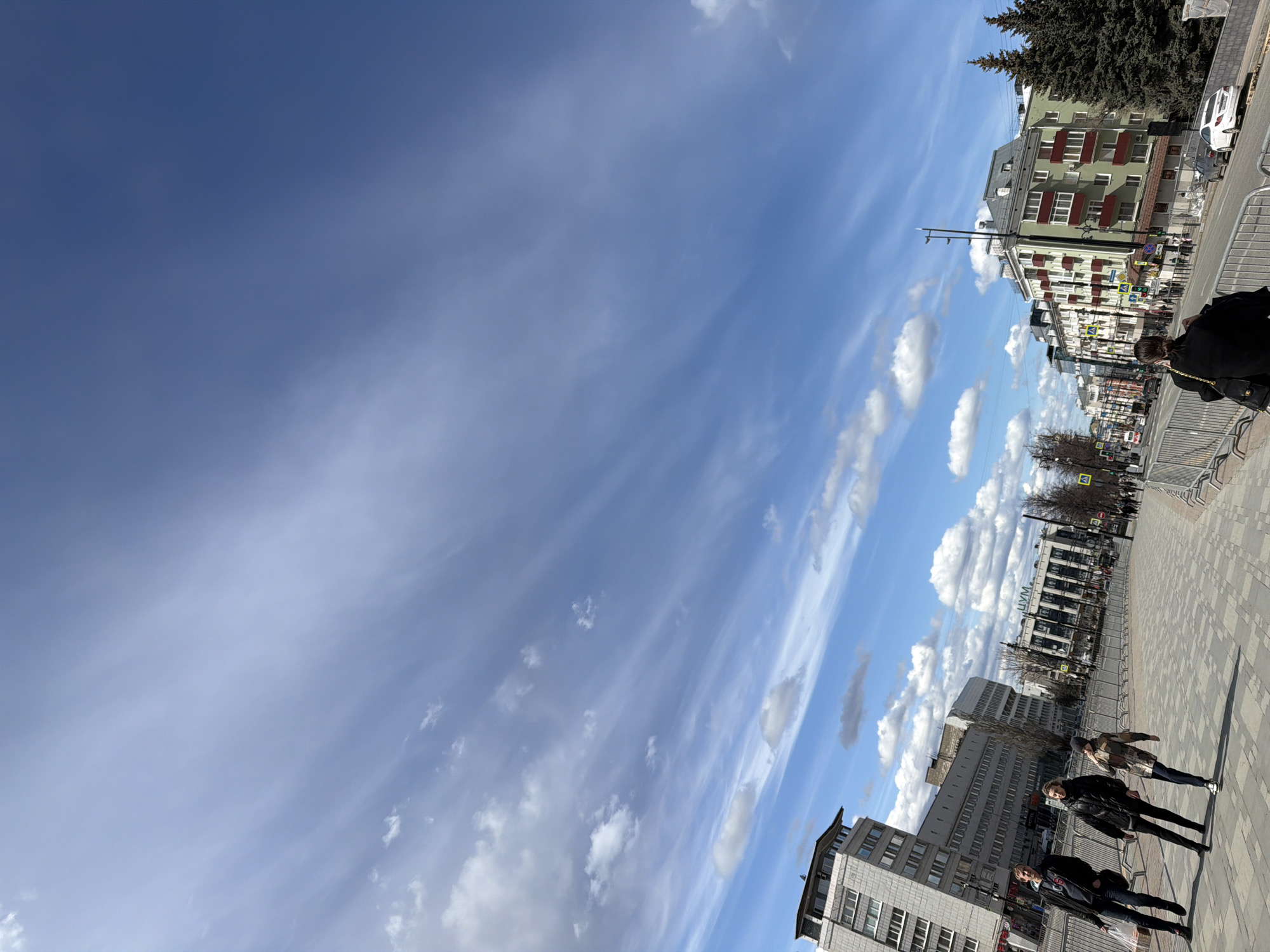 The image captures an urban street scene under a bright, clear blue sky with scattered clouds. The perspective looks down a street lined with buildings and a pedestrian area.
In the foreground and midground, several people are walking casually on a paved surface. They are dressed in coats and jackets, suggesting a cool but sunny day. Metal barriers separate the pedestrian zone from what appears to be a roadway.
On the right side of the frame, multi-story buildings line the street. The closest visible building features a green facade with white balconies and red railings. Further down the street, other buildings with grey and white exteriors are visible, including one in the distance with the Cyrillic text "ЦУМ" (TSUM) prominently displayed, indicating a department store. A large, dark evergreen tree stands tall near the top right of the frame, partially obscuring the sky.
The sky occupies a significant portion of the image, displaying a deep blue hue. Various clouds are present, ranging from wispy cirrus clouds stretching across the upper left to fluffier cumulus clouds scattered throughout the rest of the sky. The bright sunlight casts visible shadows, suggesting it is midday. The overall impression is a vibrant, ordinary day in an urban setting, consistent with Perm, Russia, as provided by the context.