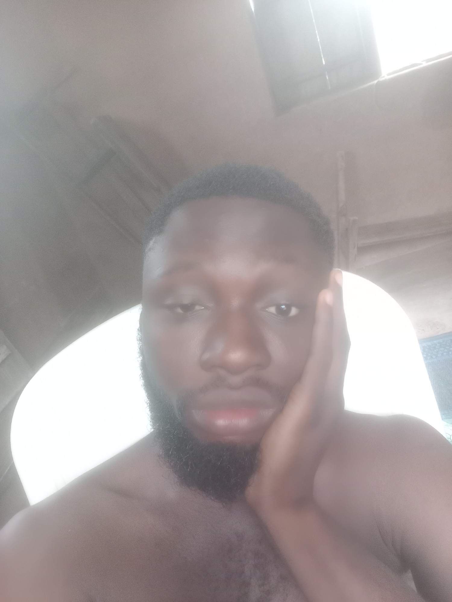 A medium close-up shot captures a Black man with dark, short hair and a full beard. His right hand is positioned against the right side of his face. The man appears to be shirtless, with a portion of his shoulder visible. The background is softly illuminated, featuring architectural elements, including what looks like part of a ceiling or roof, and an obscured bright light source. The location is assumed to be in Bende, Nigeria.