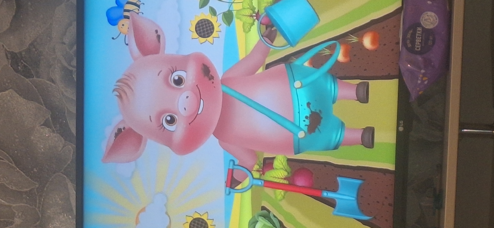 The image shows a cartoon piglet wearing blue overalls, covered in mud, holding a shovel and a watering can in a garden. The piglet has a happy expression and is surrounded by sunflowers and plants. The background depicts a bright blue sky with a sun and clouds, and a bee flying nearby. The scene is set in a garden with rows of vegetables. The lower part of the image shows the bottom of a television screen with a package of wet wipes on a shelf below. There is no information in the image that suggests its location in Bila Tserkva, Ukraine.