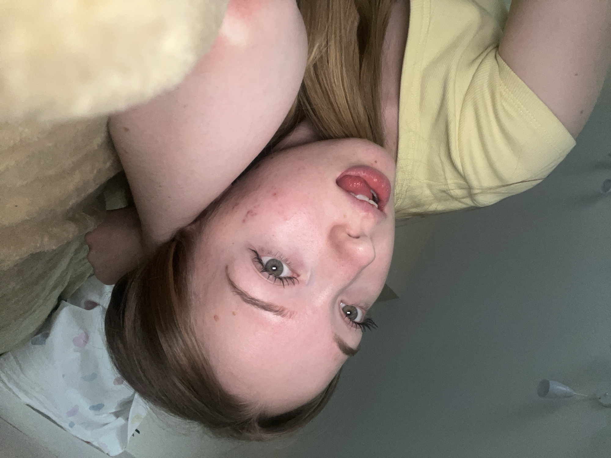 A young woman with light skin and brown hair is lying on a bed, facing the camera upside down. She is wearing a yellow short-sleeved shirt. Her mouth is slightly open, revealing her tongue. The background includes a light-colored wall, a partial view of a blanket, and a ceiling fan. It is not possible to identify the location as the city of Chaikovsky, Russia, with the given information.