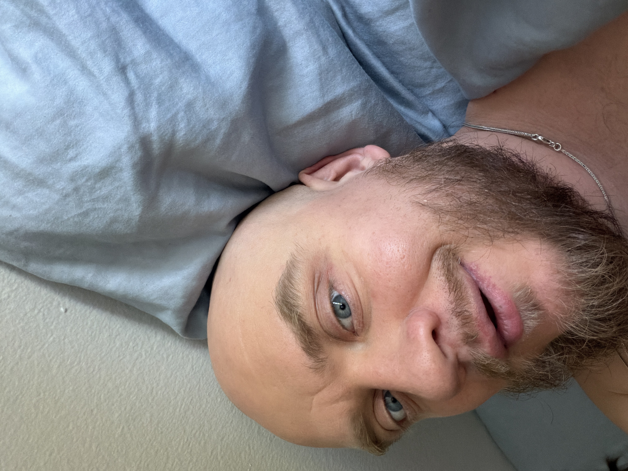 A close-up, angled portrait shows a man lying on his side. He has a shaved head, light eyebrows, blue eyes, and a beard. He wears a thin silver chain around his neck. He is lying on a pale gray or off-white surface, possibly a bed or a couch, with a light blue fabric above him. The location is unknown and cannot be determined from the image; therefore, the location of Morton Grove, United States of America, cannot be verified.