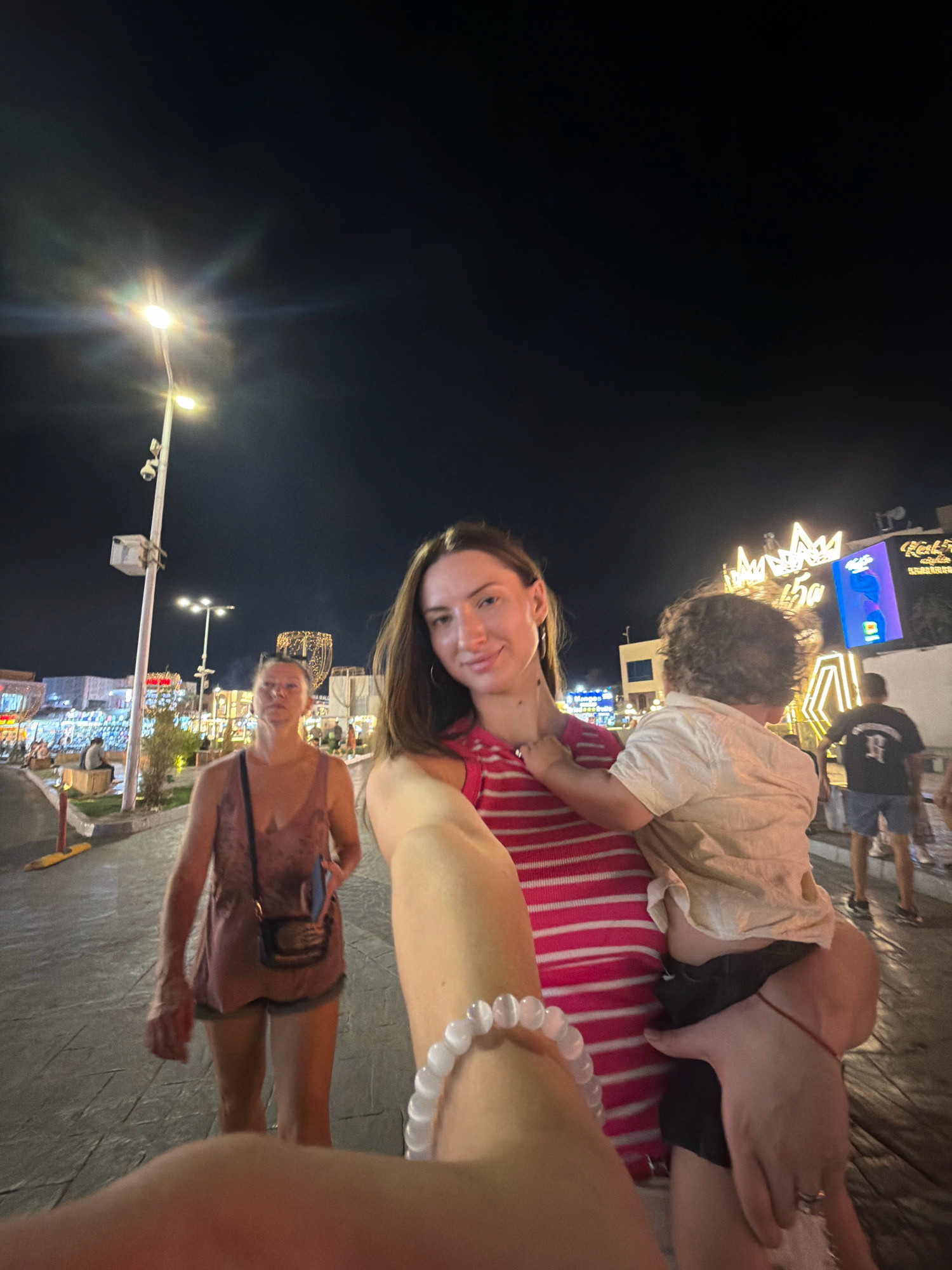 A woman is taking a selfie while holding a young child in her arms at night in a pedestrian area of Sharm El Sheikh, Egypt. She is wearing a pink and white striped top and the child is in a light-colored shirt. In the background, another woman is walking away from the camera, wearing a pink tank top and shorts. The scene is lit by streetlights and illuminated buildings, suggesting a commercial or entertainment district.