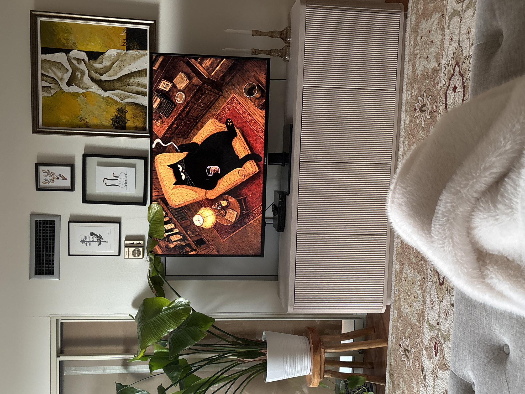 The image depicts a cozy living room scene, likely within a residence in Richmond Hill, Canada, given the context provided. The focal point is a large television screen displaying a whimsical animated image of a fat black cat lounging in an armchair, smoking a cigarette and holding a drink. The cat appears content and relaxed.
Above the television, the wall is adorned with several framed pictures. To the left, there's a vent and a framed artwork of a bird. Next to it are smaller frames featuring a deer head illustration and a minimalist depiction of three birds. To the right of these, a larger, abstract painting with prominent yellow and white textures hangs.
The television is placed on a light-colored media console with a fluted design. To the left of the television, a large potted plant with broad green leaves is situated on a rustic wooden stool. In the foreground, a soft, plush cream-colored blanket is draped over a tufted grey sofa, suggesting a comfortable viewing experience.
The flooring is covered by a patterned rug with intricate designs in muted tones of brown, beige, and red. The overall ambiance of the room is one of warmth and comfort, with a touch of playful personality evident in the choice of artwork. The lighting appears to be a mix of ambient room light and the illumination from the television screen.