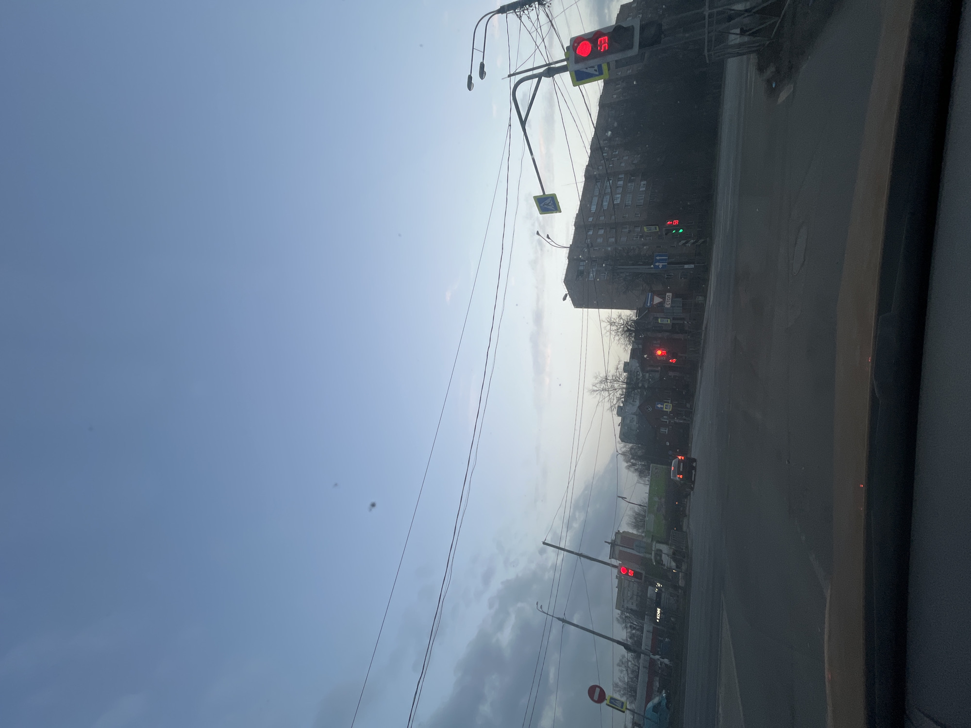 The image, taken from inside a vehicle, shows a street scene in Klin, Russia. The sky is overcast. Several traffic lights are visible, most displaying red. Buildings line the street, including what appears to be a large apartment block. A car is visible in the distance. Power lines crisscross the sky. The road surface is asphalt.