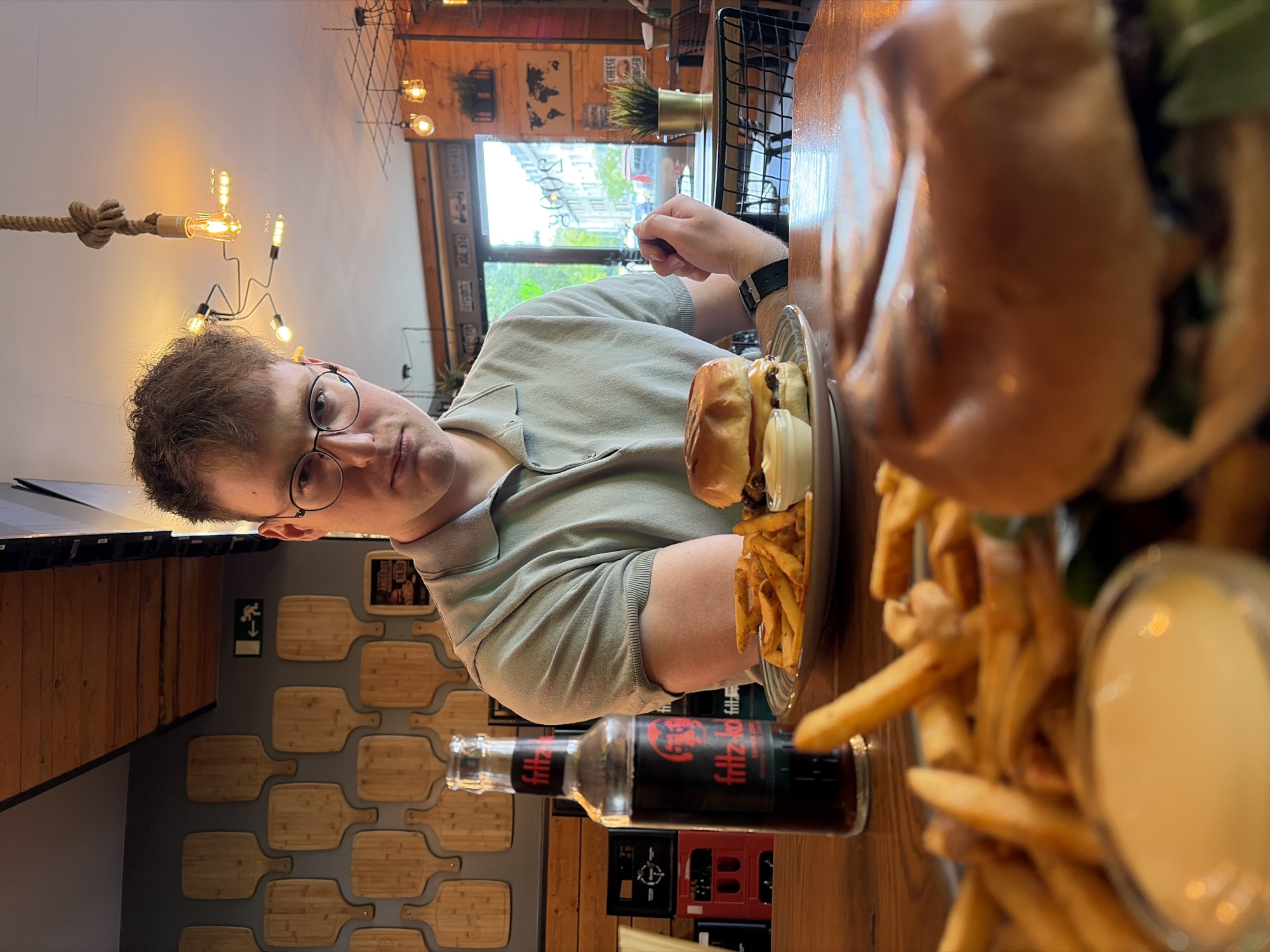 A young man with light brown hair and glasses, wearing a light-colored polo shirt, sits at a wooden table in a restaurant, looking directly at the camera. He has a neutral expression. In front of him, on the table, are several food items: a plate holding a burger with a side of French fries and a small dish of sauce. Another, larger burger, also accompanied by fries and sauce, is partially visible and out of focus in the foreground. A glass bottle of "Fritz-kola" stands on the table.
The restaurant's interior is warmly lit by decorative industrial-style light fixtures, including one with a knotted rope design. The background features a large window revealing an outdoor urban street scene, indicating it is daytime. The walls are adorned with various decorations, including a prominent section covered in wooden cutting board shapes arranged in a grid. Other wall decor includes framed pictures or posters, and license plate-like items reading "J0C 870" and "PIS 200". A sign mentioning "CHEESE BURGER" is also visible. A green emergency exit sign with a downward arrow is on one wall. The setting suggests a casual dining experience, consistent with a burger joint in Essen, Germany.