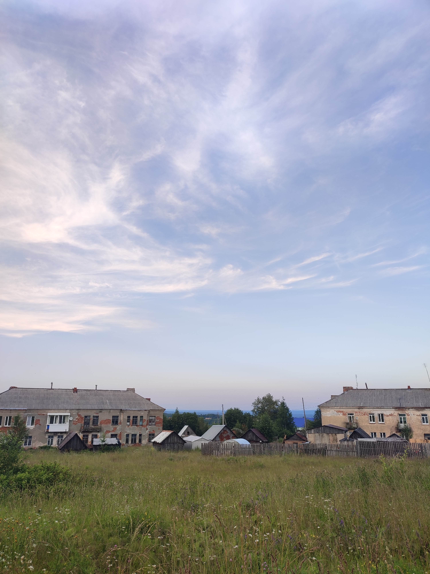 The image depicts a rural landscape with two weathered, two-story apartment buildings flanking a field of tall grass and wildflowers. The buildings, constructed with brick and featuring corrugated metal roofs, show signs of age and disrepair. Small, rustic wooden sheds and a wooden fence are visible between and in front of the buildings. The sky above is a soft blue, filled with wispy, white clouds. In the distance, a line of trees and a body of water are faintly discernible, suggesting the presence of a lake or river. The overall impression is one of a quiet, somewhat dilapidated rural settlement, possibly in the city of Gremyachinsk, Russia, given the provided context. There are no people visible in the image.